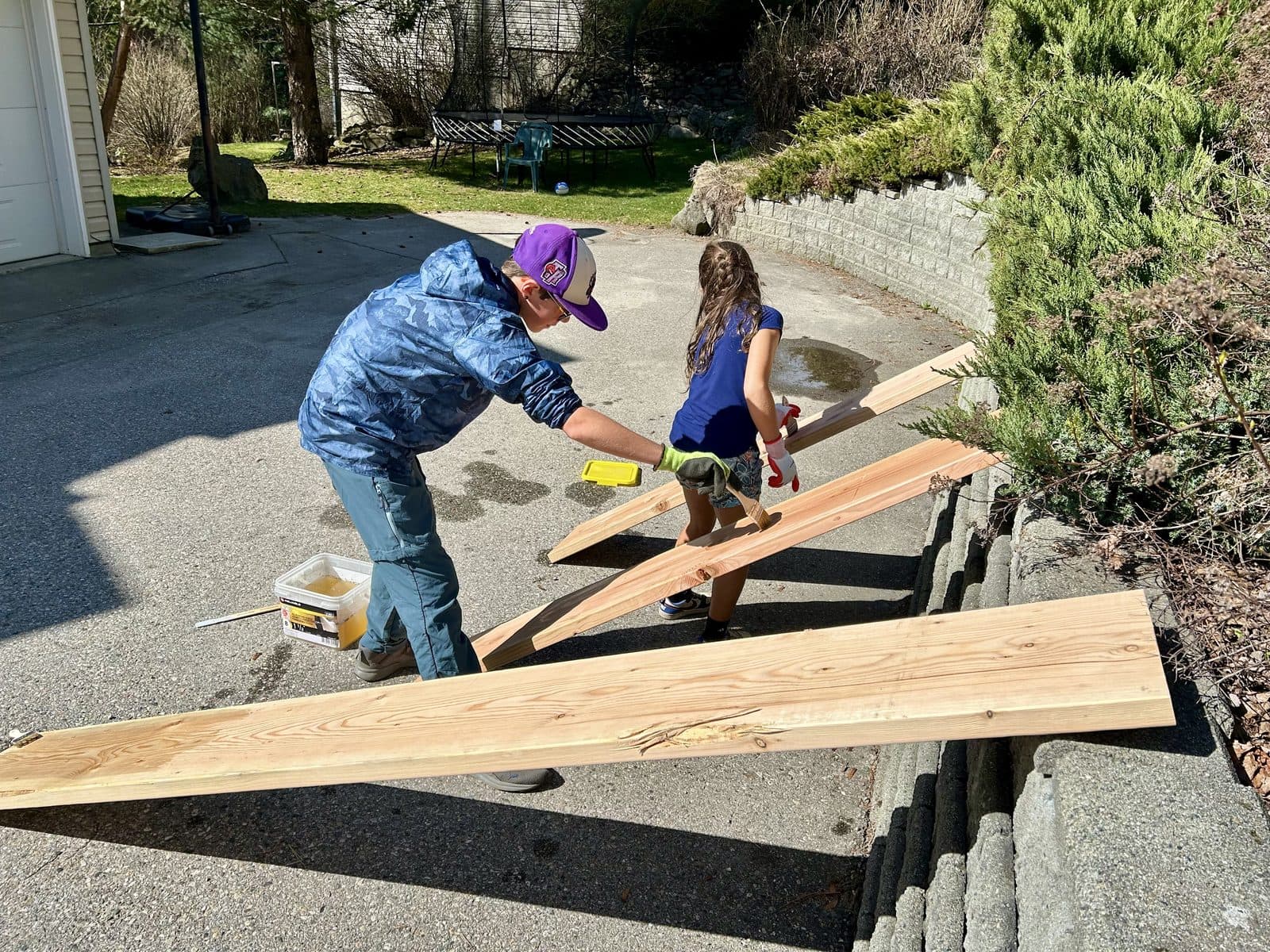 Two kids working together on a wooden building project in the driveway, drill and lumber in hand