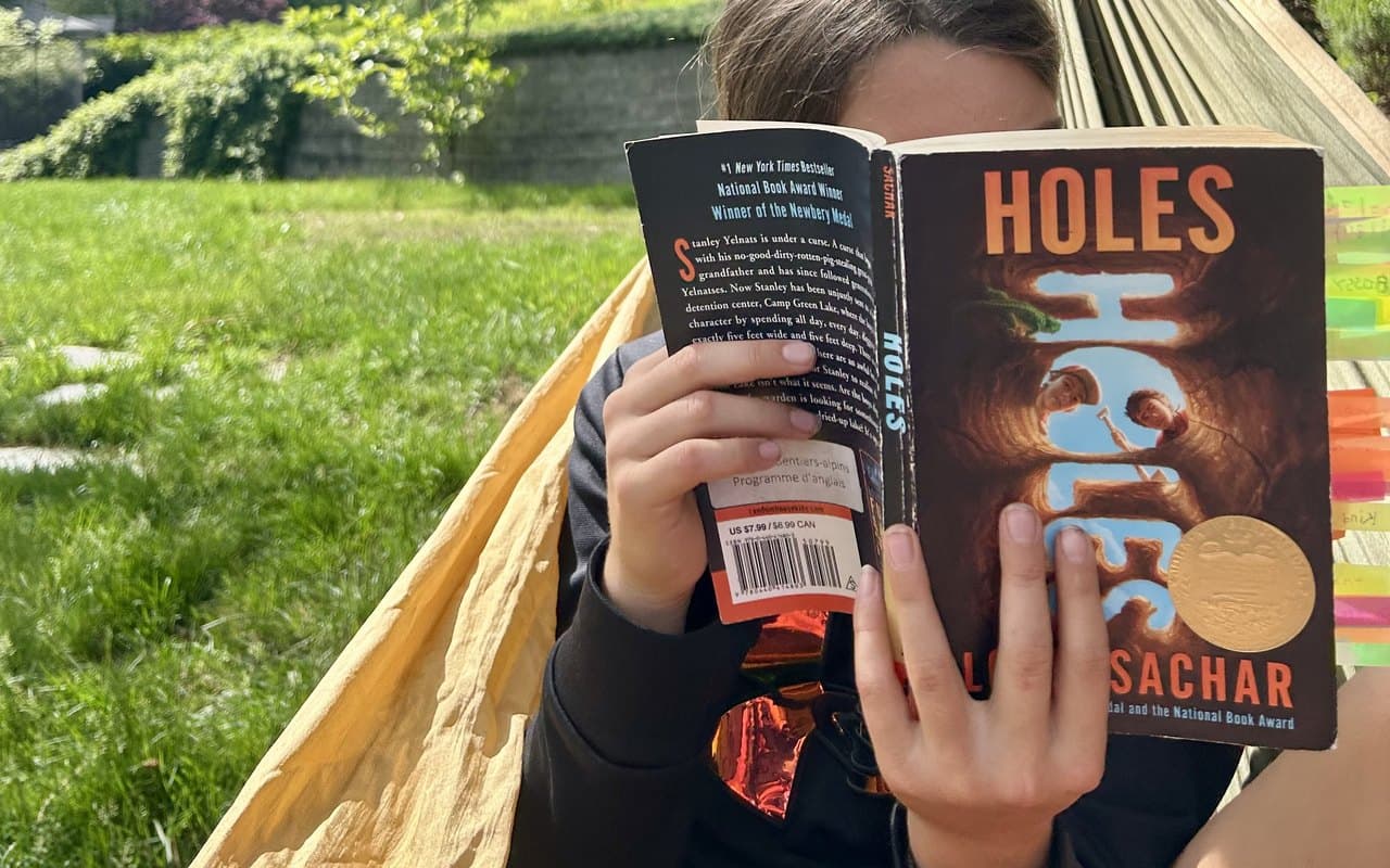 A child reading the novel Holes in a backyard hammock on a sunny day, deep in a book with a green yard and forest behind