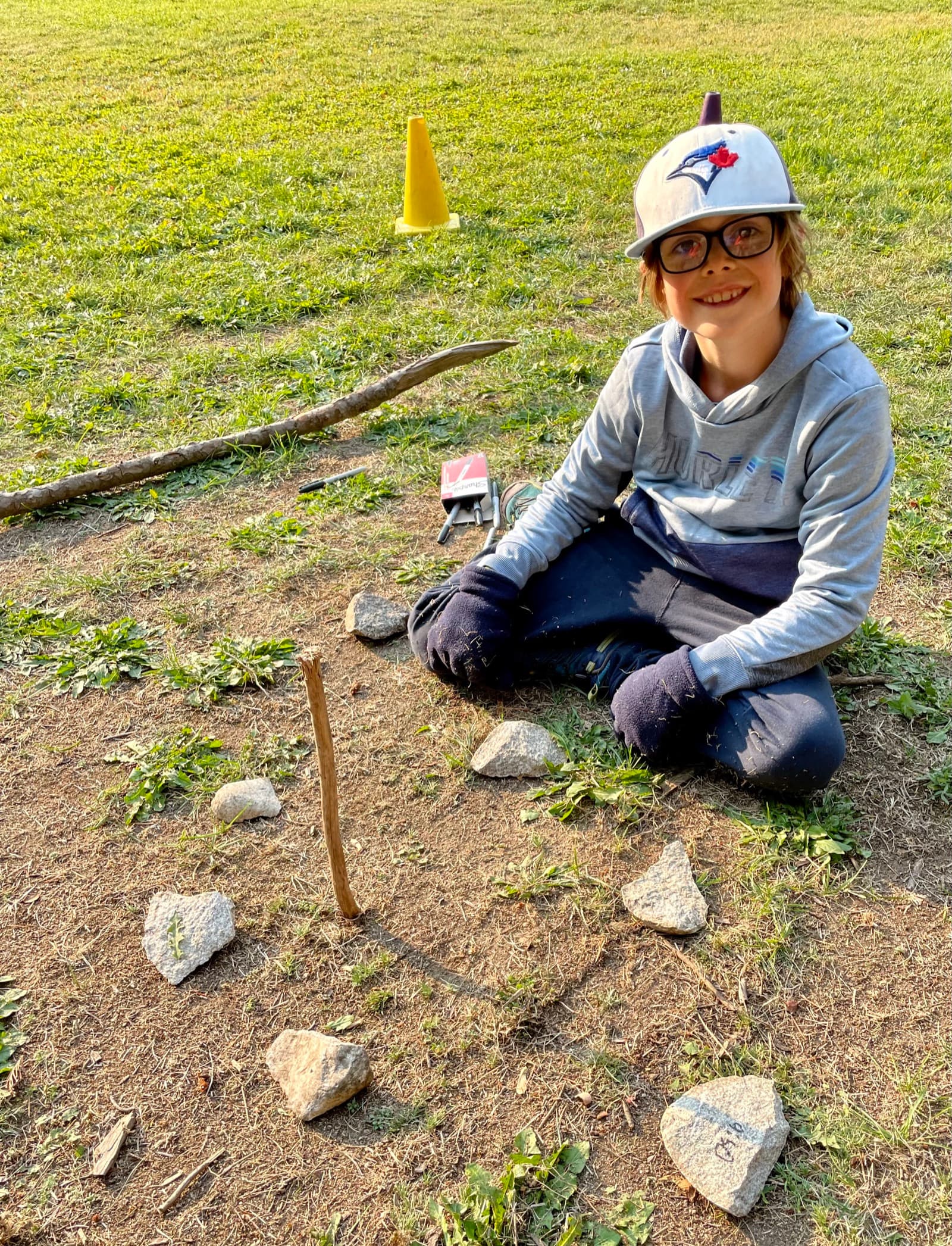 Boy kneeling next to a homemade sundial made from a stick and rocks arranged in a curve on the grass
