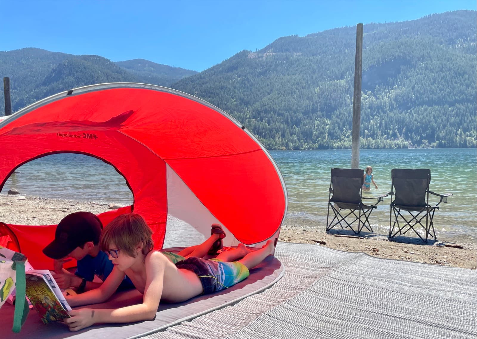 Two kids lounging in a red beach shelter on a lazy summer afternoon by the lake, one reading a comic, with nothing scheduled and nowhere to be