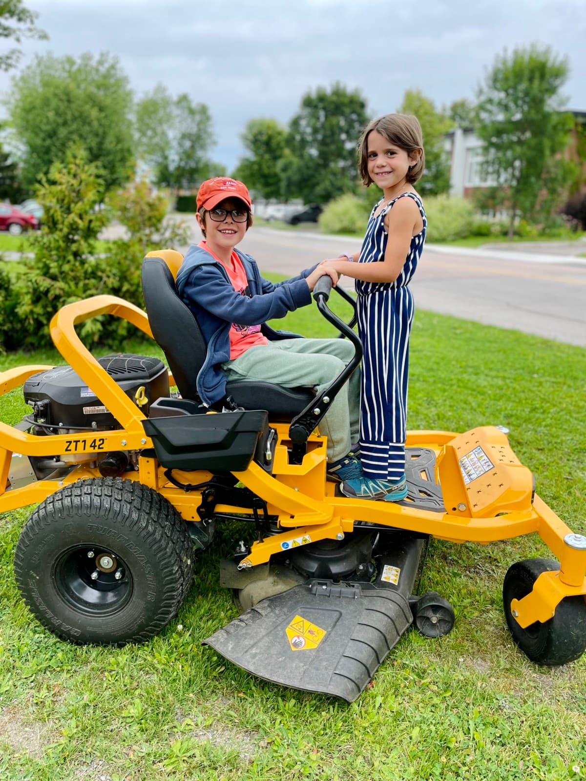 Two kids learning to operate a riding lawn mower together on the front yard