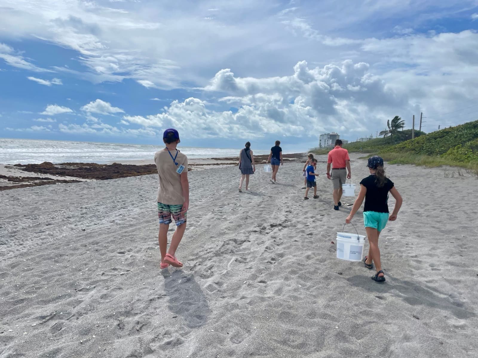 Kids and adults carrying white buckets along a windy Florida beach during a beach cleanup, seaweed on the tideline and dune grass to the right