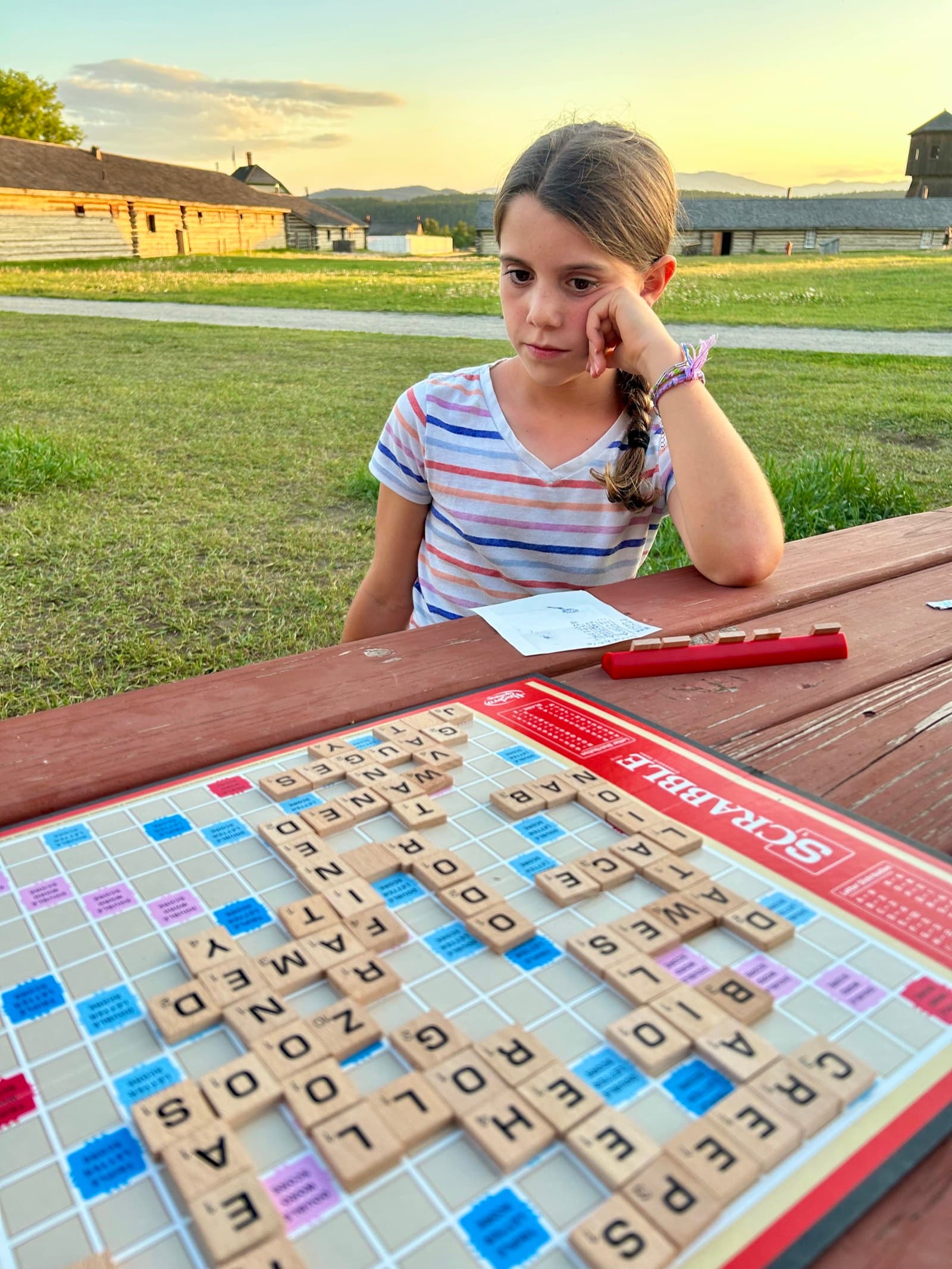 Girl studying a Scrabble board at an outdoor picnic table during golden hour, chin on hand, deep in thought
