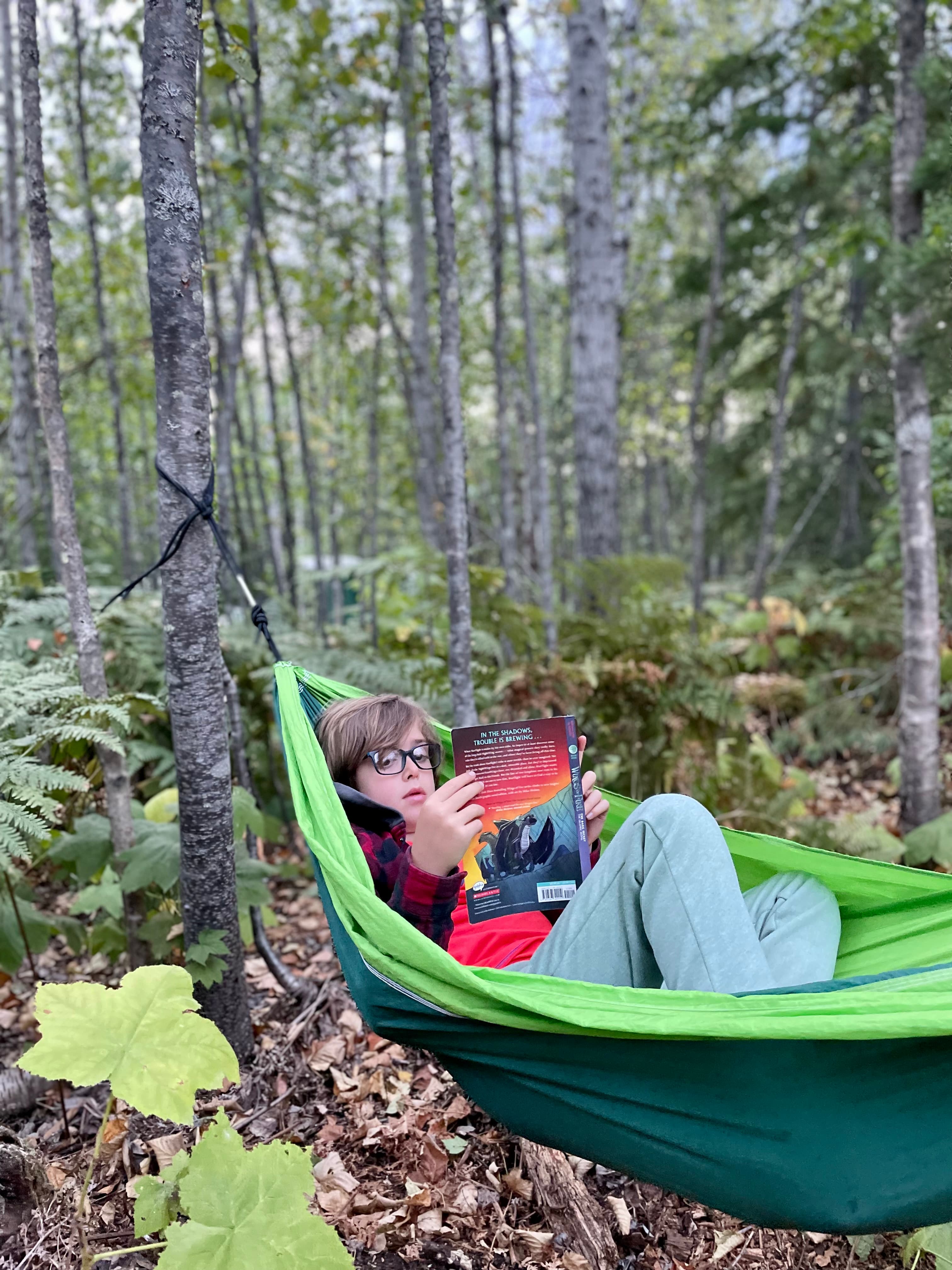 Kid reading a book in a hammock strung between trees in the forest