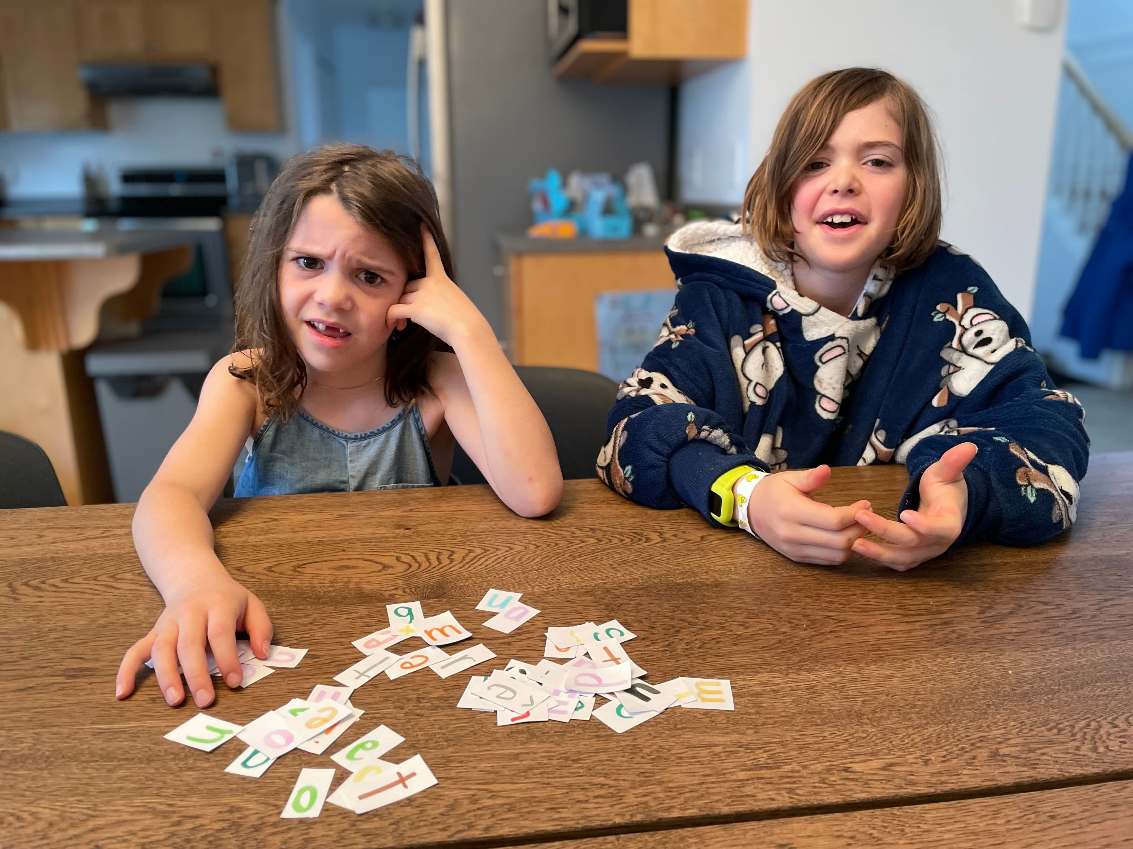 Two kids at the kitchen table with letter tiles, one frustrated, the other animatedly explaining