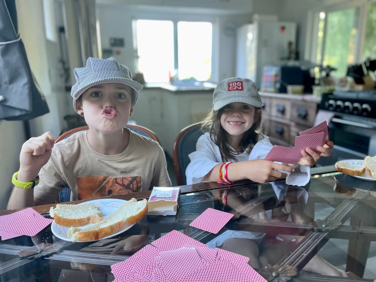 Two kids sitting at a kitchen table with train conductor hats, playing a card game and making strategic choices