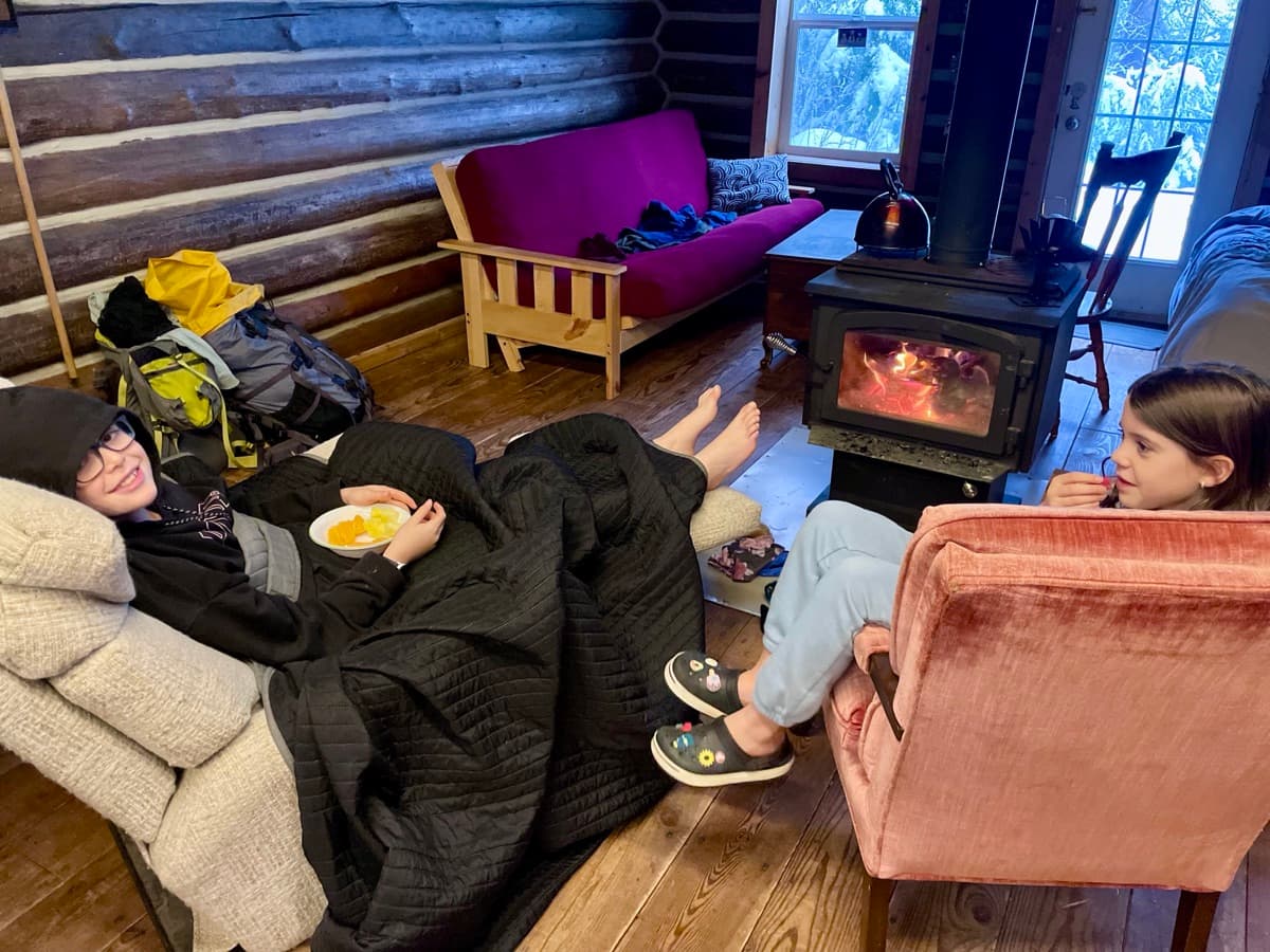 Both kids relaxing on a couch by a wood stove with bowls of snacks, the kind of unstructured cozy afternoon that does the most quiet deschooling work