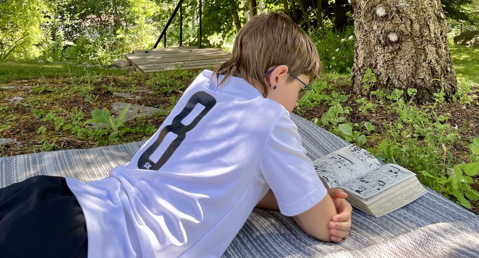 Zach lying on a picnic blanket under tall trees, reading at his own pace in dappled afternoon light, the slow shape of deschooling