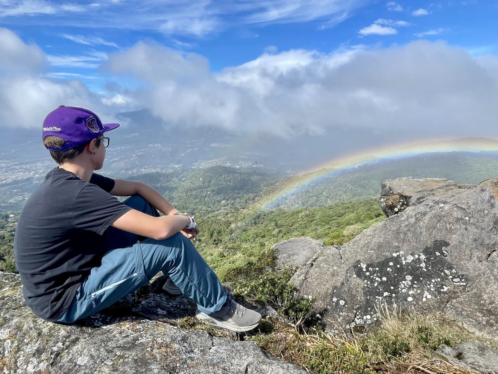 My son sitting on a mountaintop looking out at a rainbow over the valley, the quiet reflection of deschooling