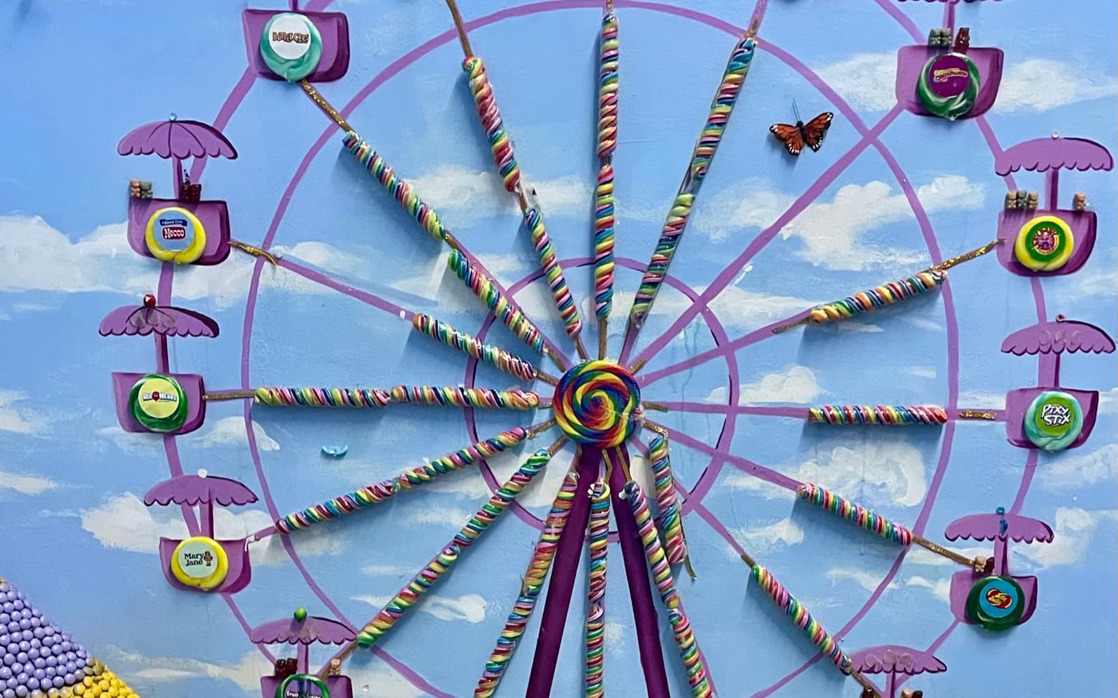 A wall-sized Ferris wheel built from candy: rainbow lollipop spokes, purple umbrella-topped cars each holding a different sweet, painted against a blue sky, the kind of ride a kid could dream up for their own park