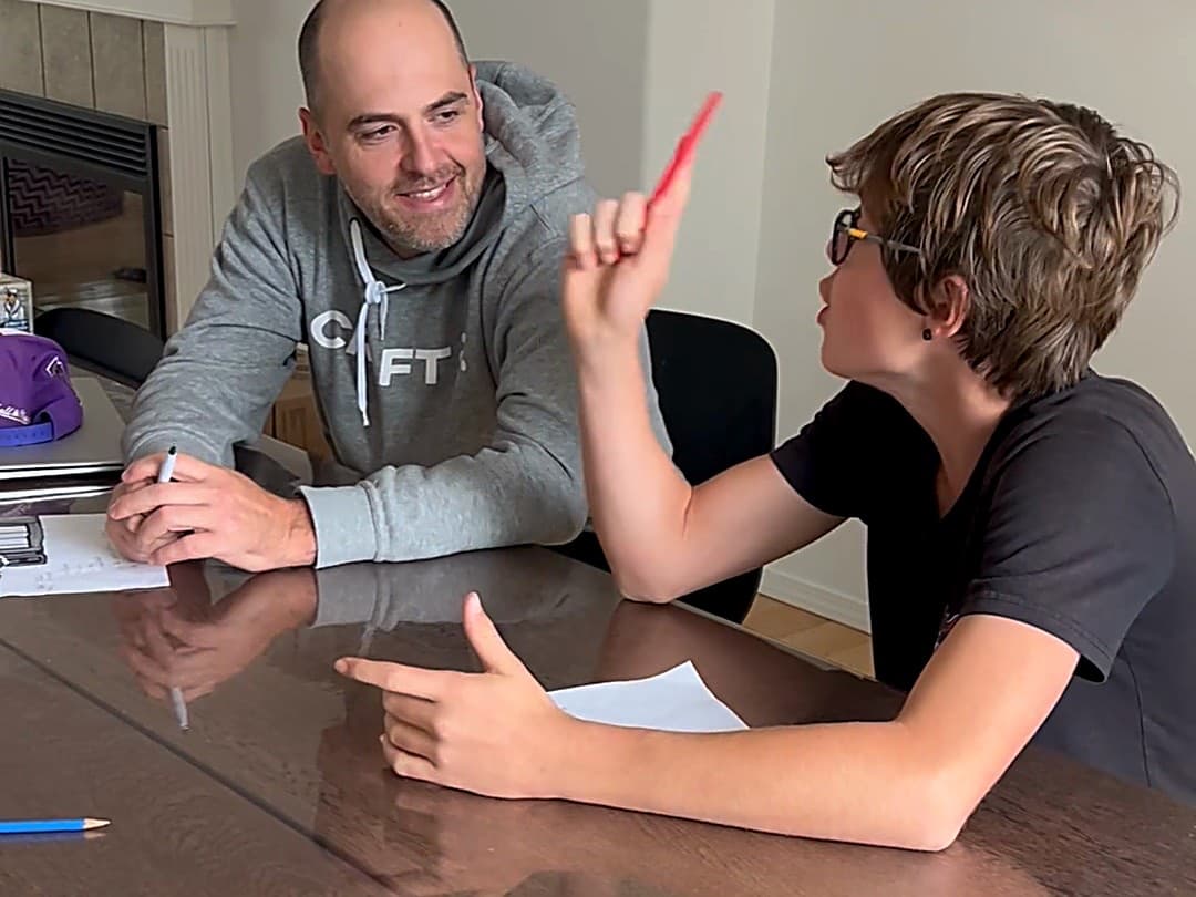 A boy in glasses raising a red pencil as he makes his point at the dining table while his dad smiles and listens, written arguments on the table between them