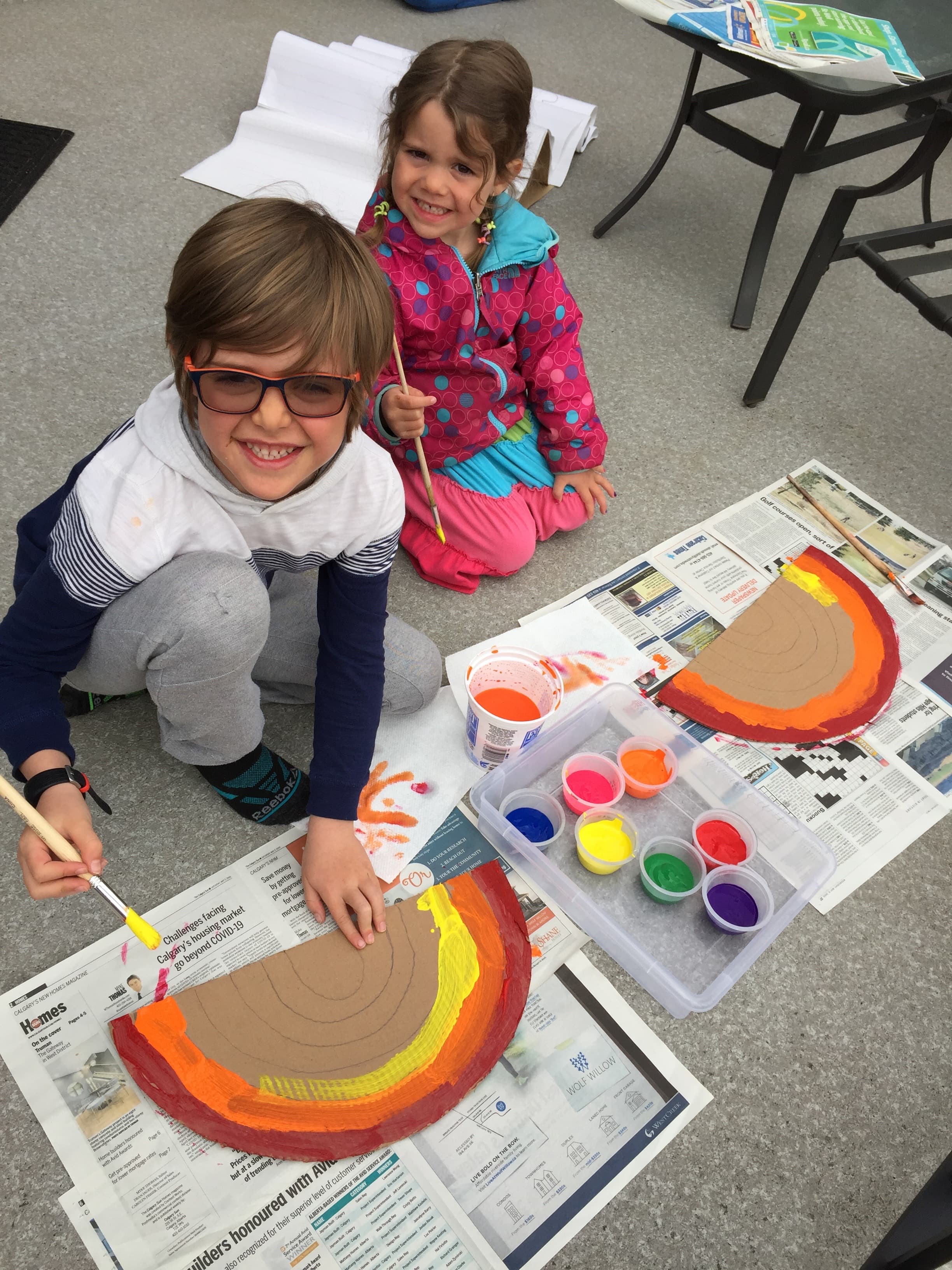 Two kids sitting on the floor painting bright cardboard rainbows with pots of colourful paint spread around them