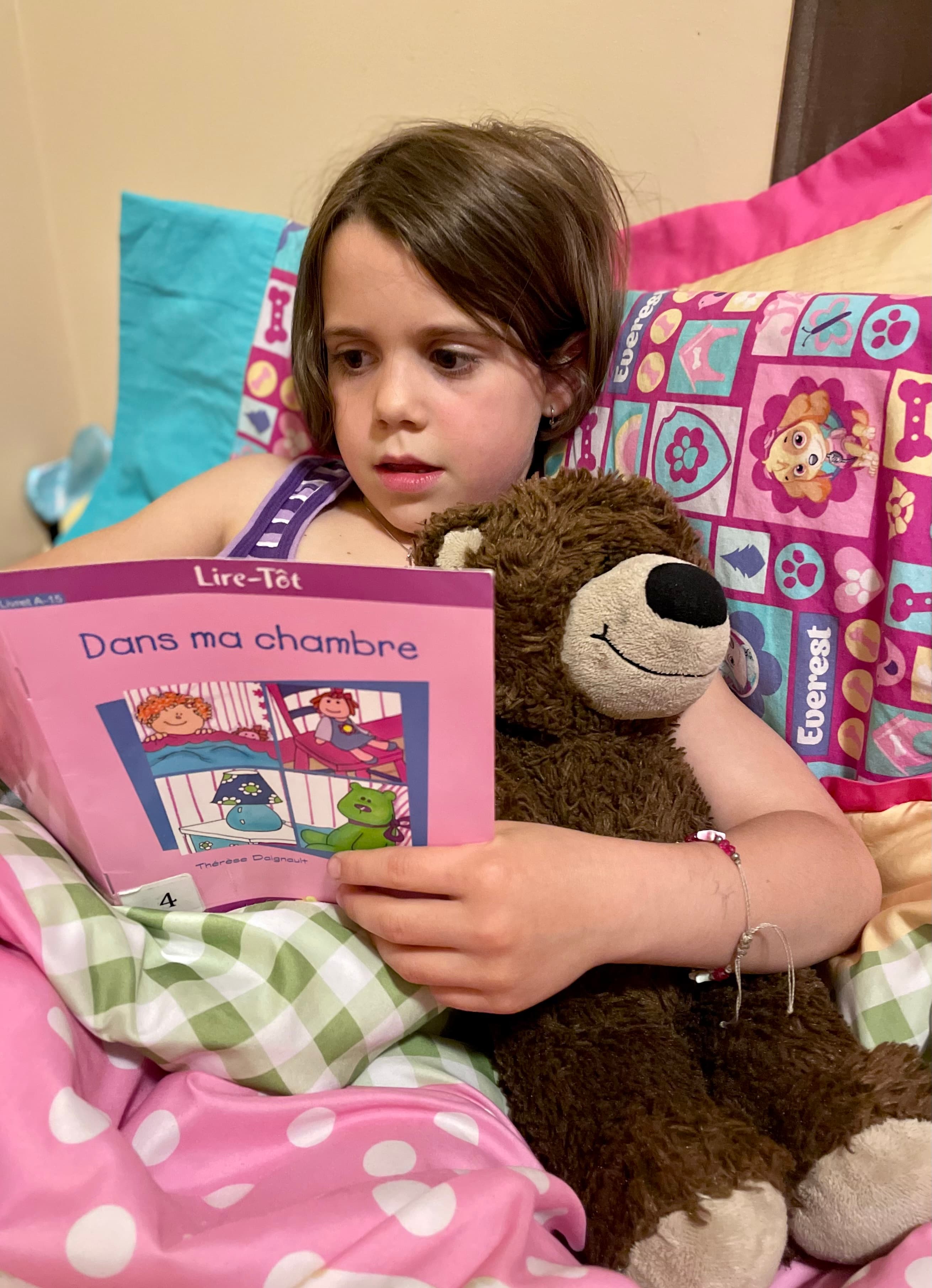 Girl reading a picture book in bed snuggled up with a stuffed bear, the kind of quiet evening reading that builds lifelong readers