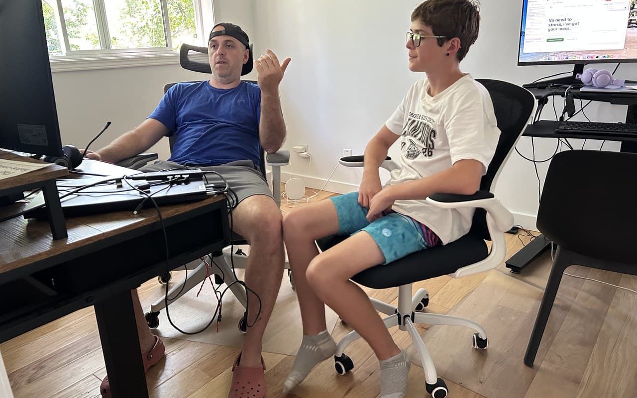 A parent working at a home-office desk with monitors while their tween sits alongside in an office chair, homeschooling and working from home side by side
