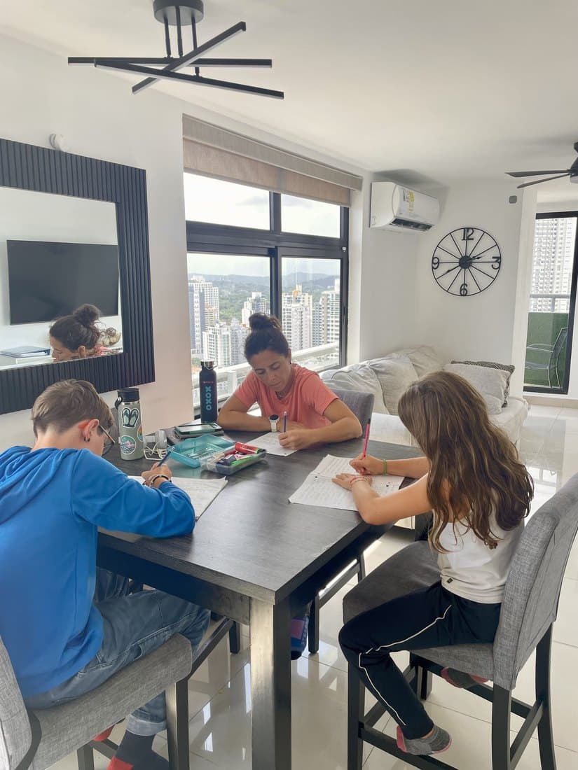 A parent and two kids all working at the same table, each on their own task, in a bright apartment