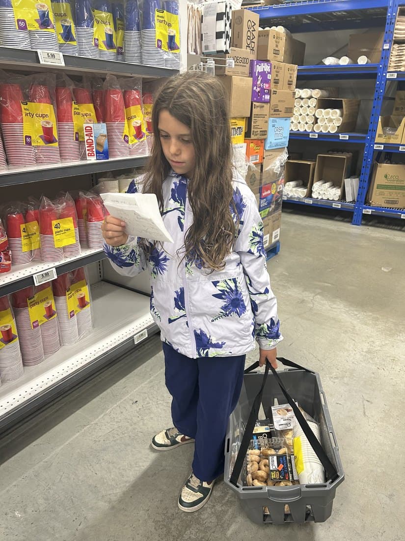 A child reading her own handwritten grocery list while carrying a basket of groceries in a store, with shelf prices visible beside her