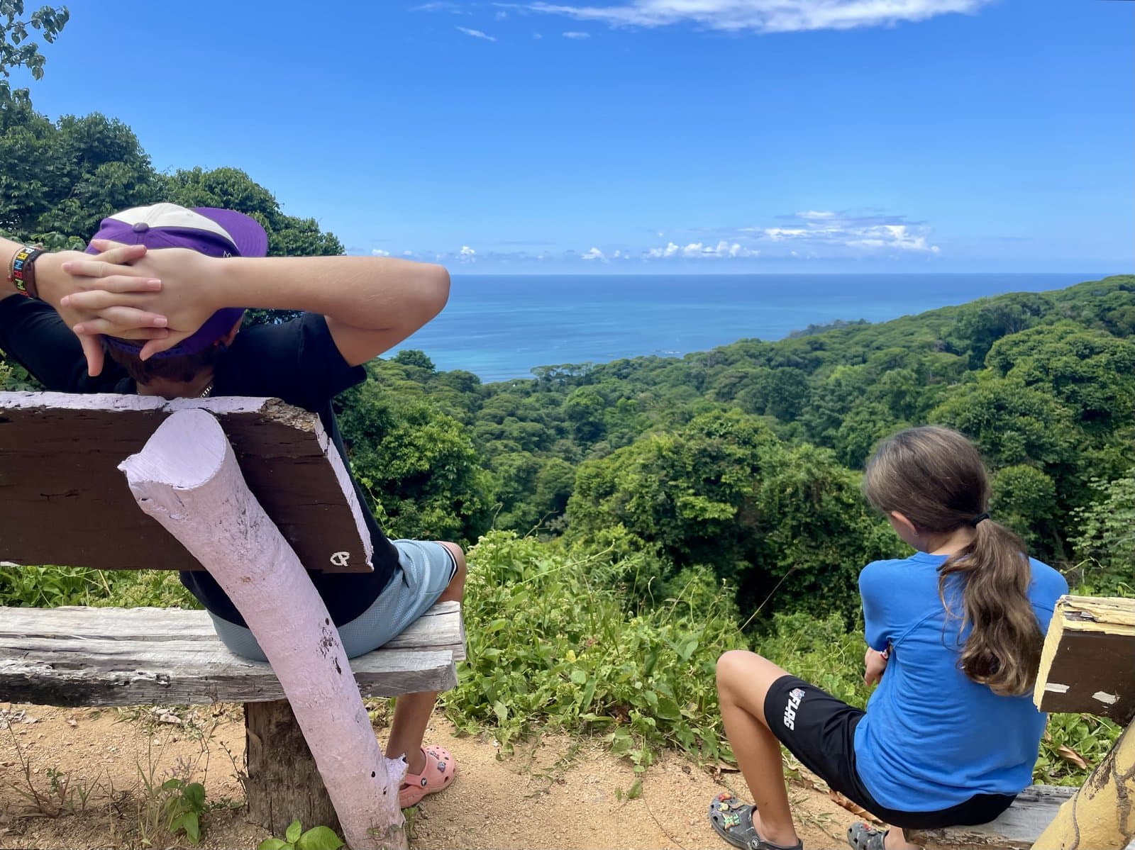 Two kids sitting on a bench at a jungle viewpoint overlooking the ocean, a quiet moment of calm