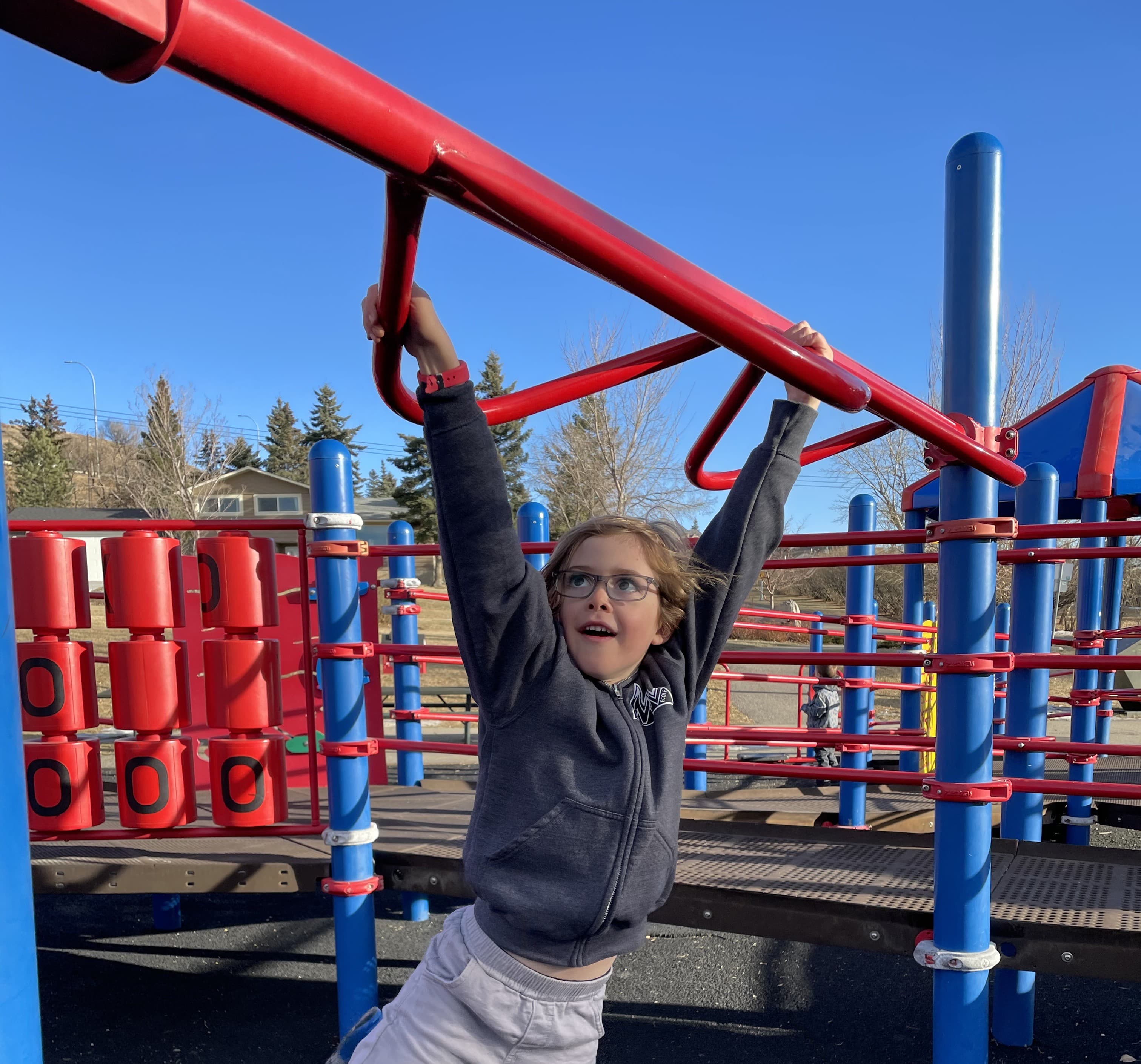 Young boy hanging from red and blue playground climbing bars against a bright blue sky, full of first-grade energy
