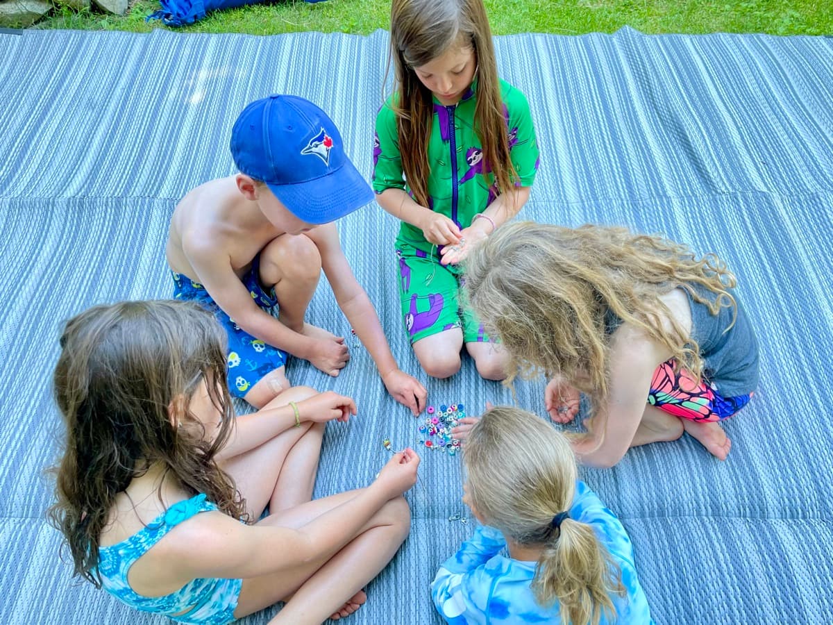 Four kids sitting on a striped blanket making bracelets together with no adult in sight, a quiet kid-led friendship moment