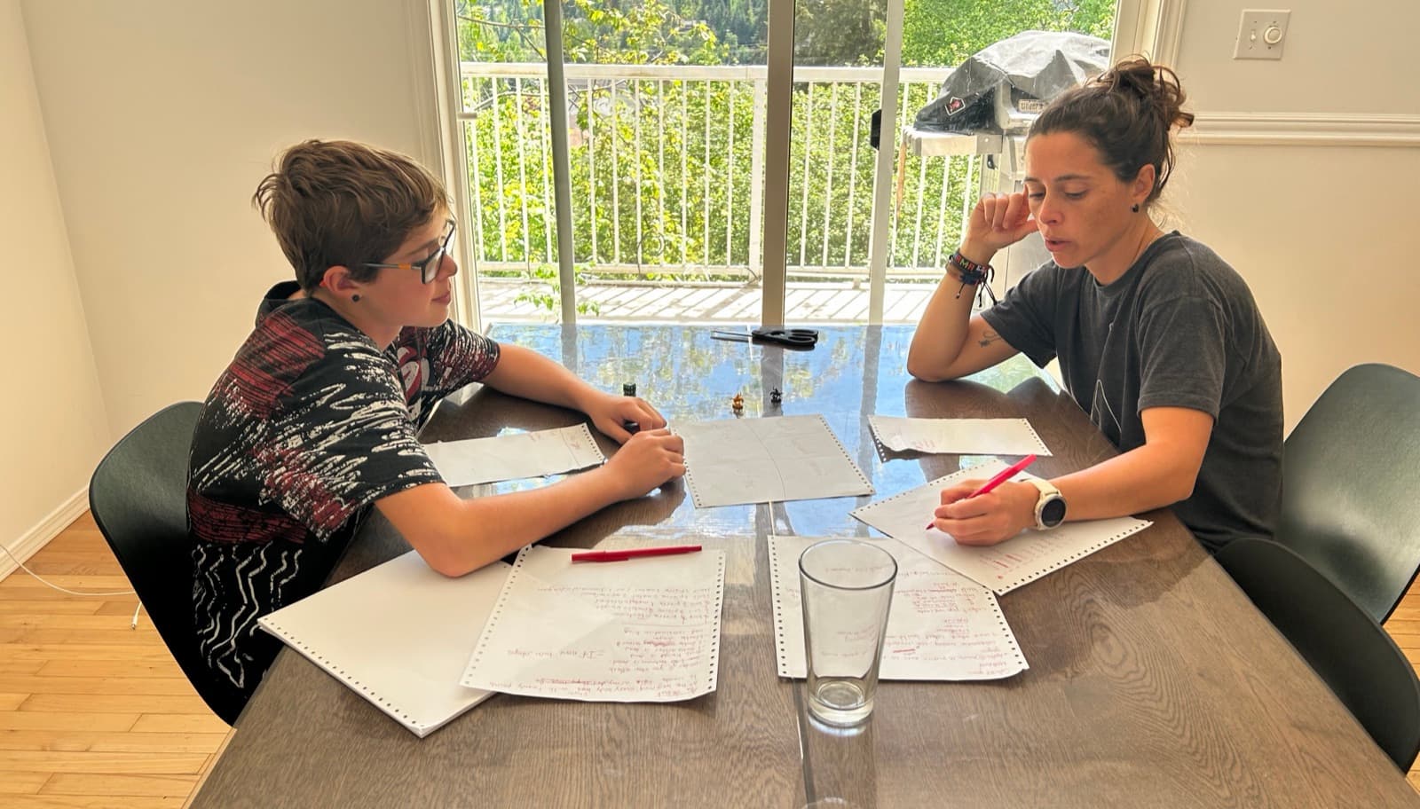 Mom and son working through math at the kitchen table together on a regular homeschool morning, big window with trees behind them