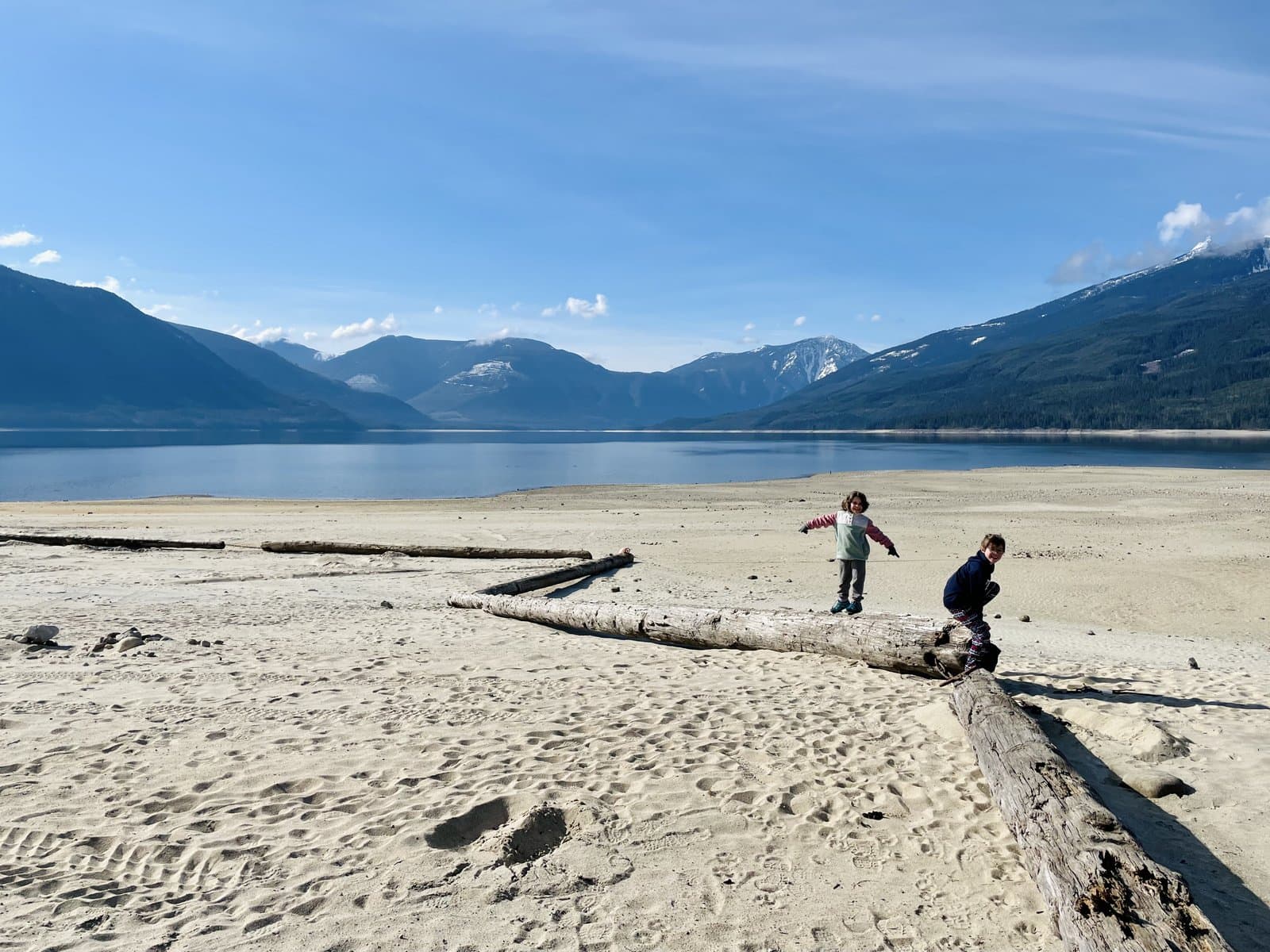 Two kids balancing on a driftwood log on a sandy lakeshore with mountains and blue sky behind them