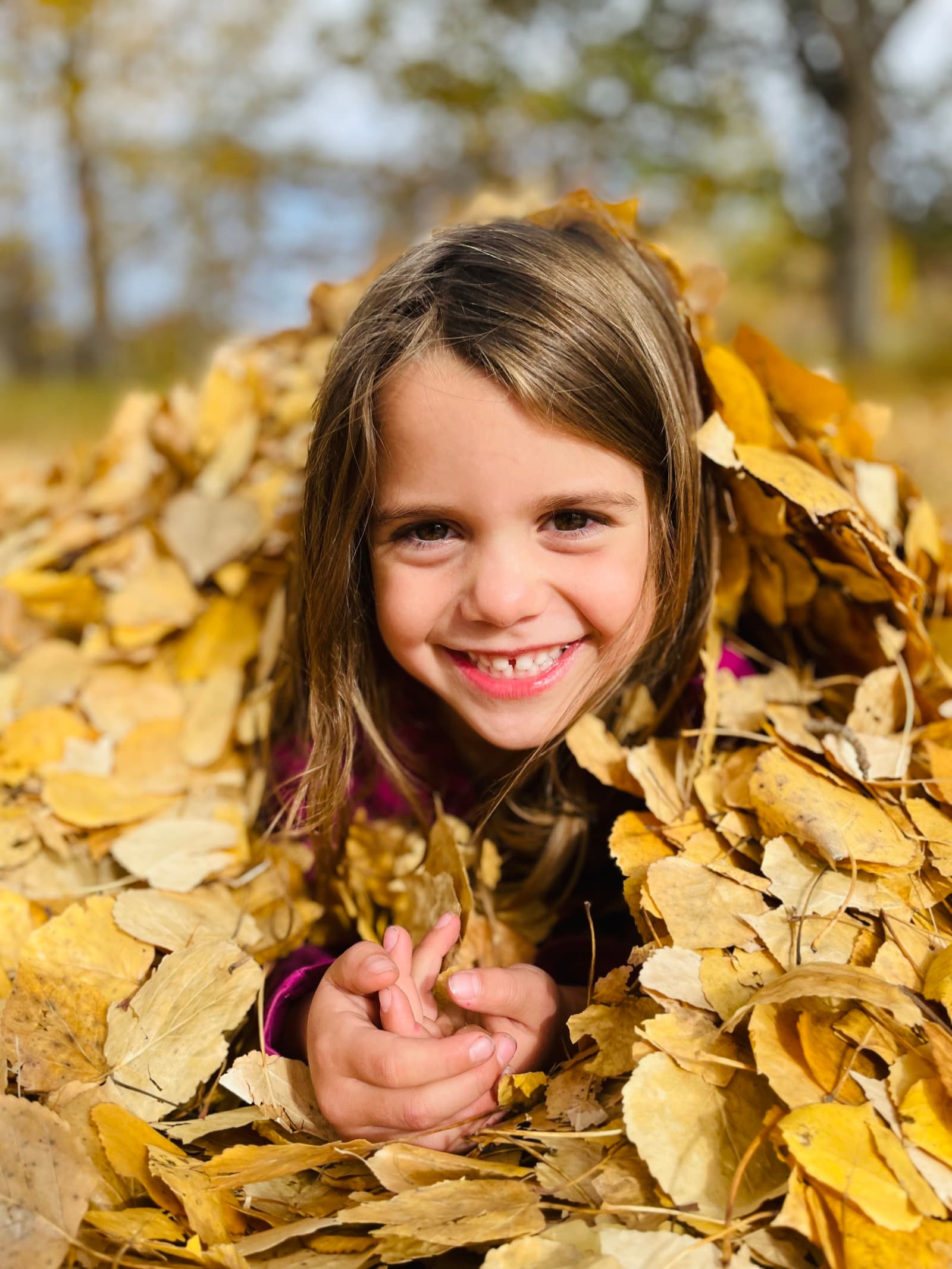 Young girl peeking out from a pile of golden fall leaves with a huge grin