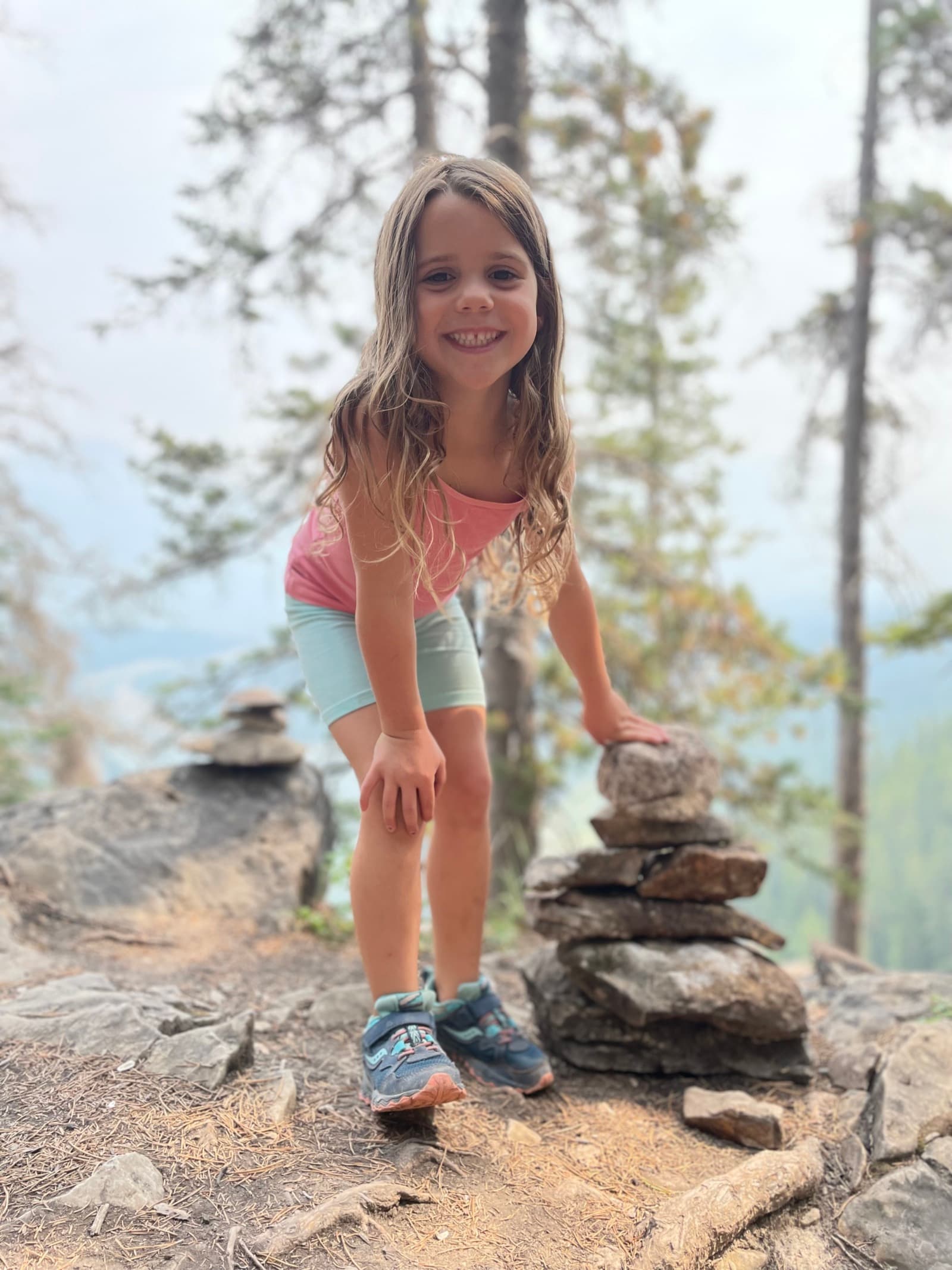 Young girl smiling next to a rock cairn she built on a mountain hiking trail
