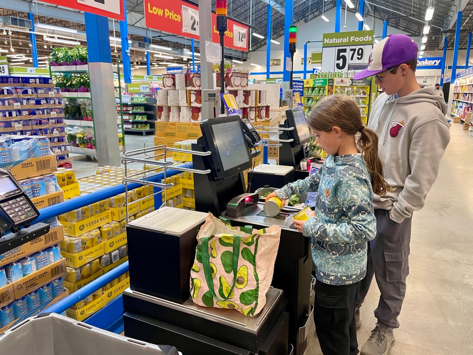 Julia scanning groceries at a self-checkout while Zach watches, handling the full transaction independently
