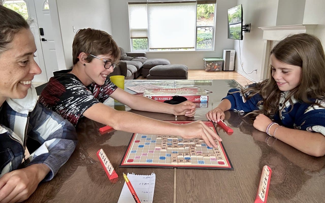 A parent and two kids gathered around the dining table playing Scrabble together, smiling, in a bright living room
