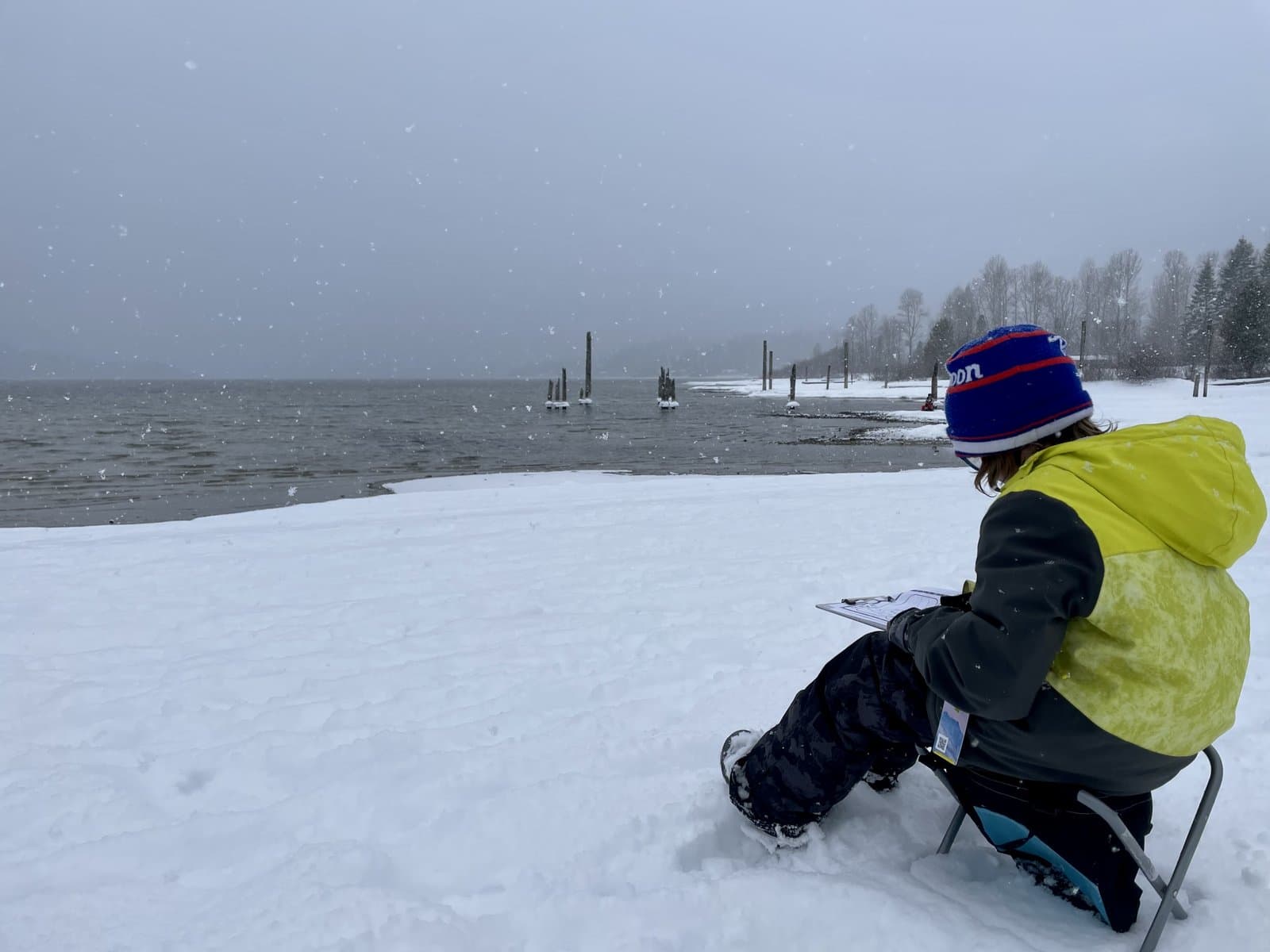 Kid sitting on a folding chair by a snowy lakeshore, drawing on a clipboard while snow falls around them, the literal practice of nature journaling