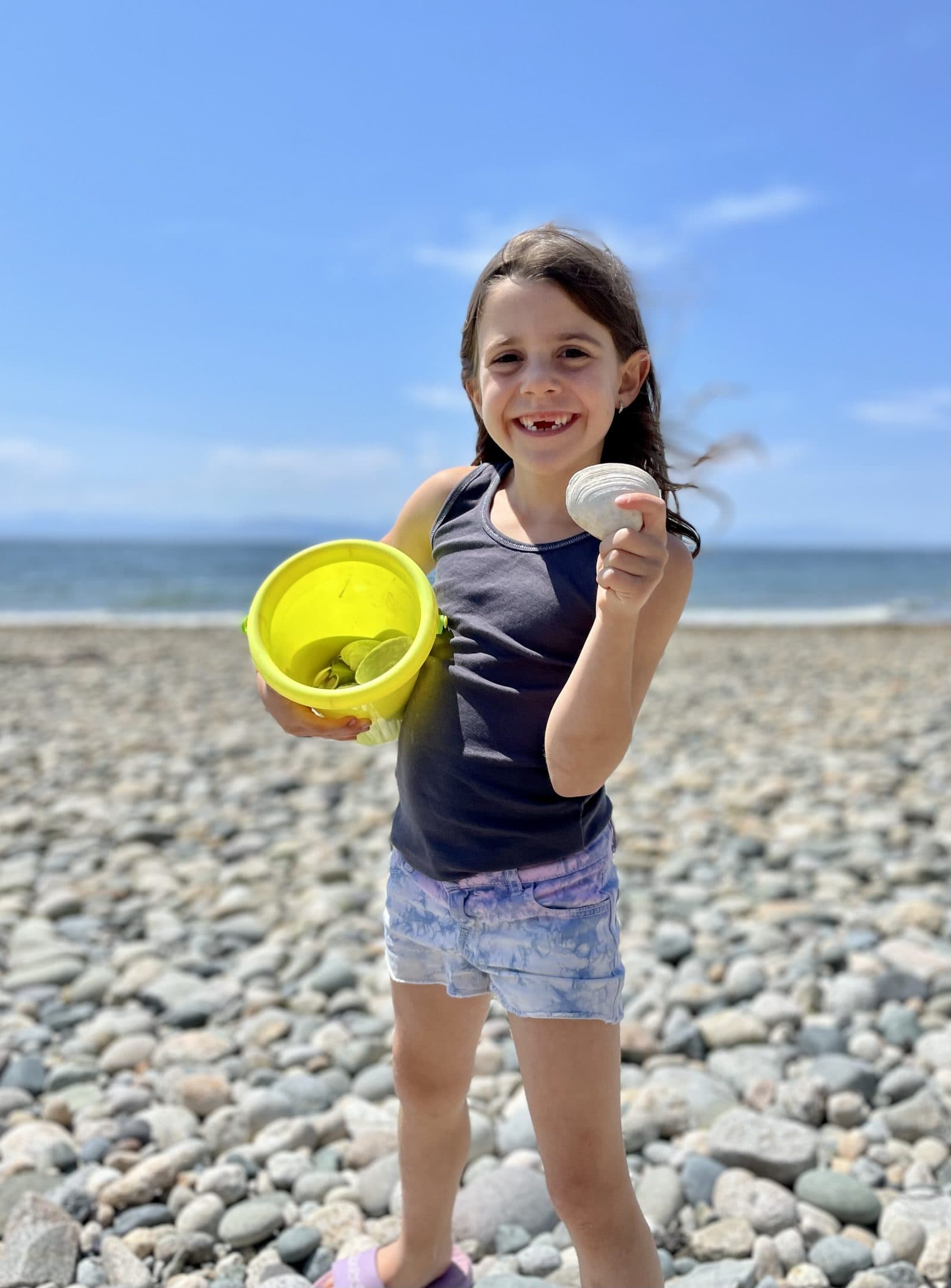 Kid on a stony beach examining a single shell from her collection bucket, the kind of close-up observation that fuels a nature journal entry