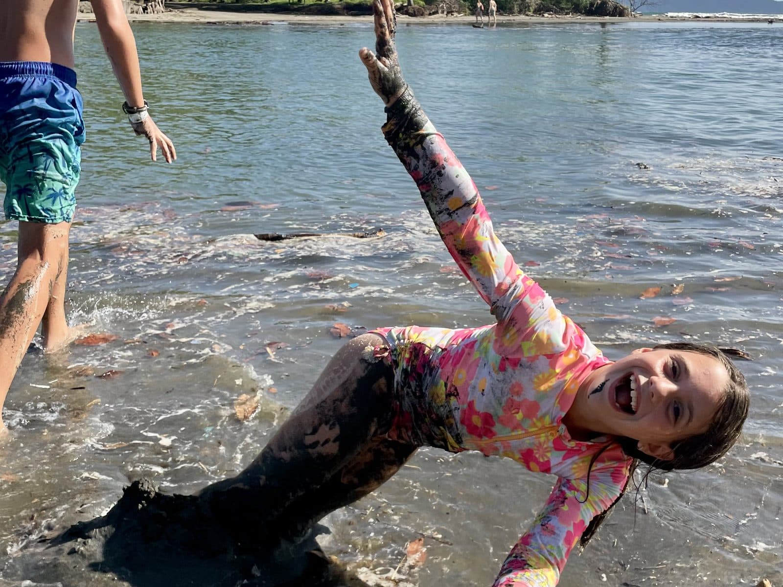 A laughing child sunk deep in soft beach mud with one muddy arm raised, testing whether the mud would hold her weight, while another child wades nearby