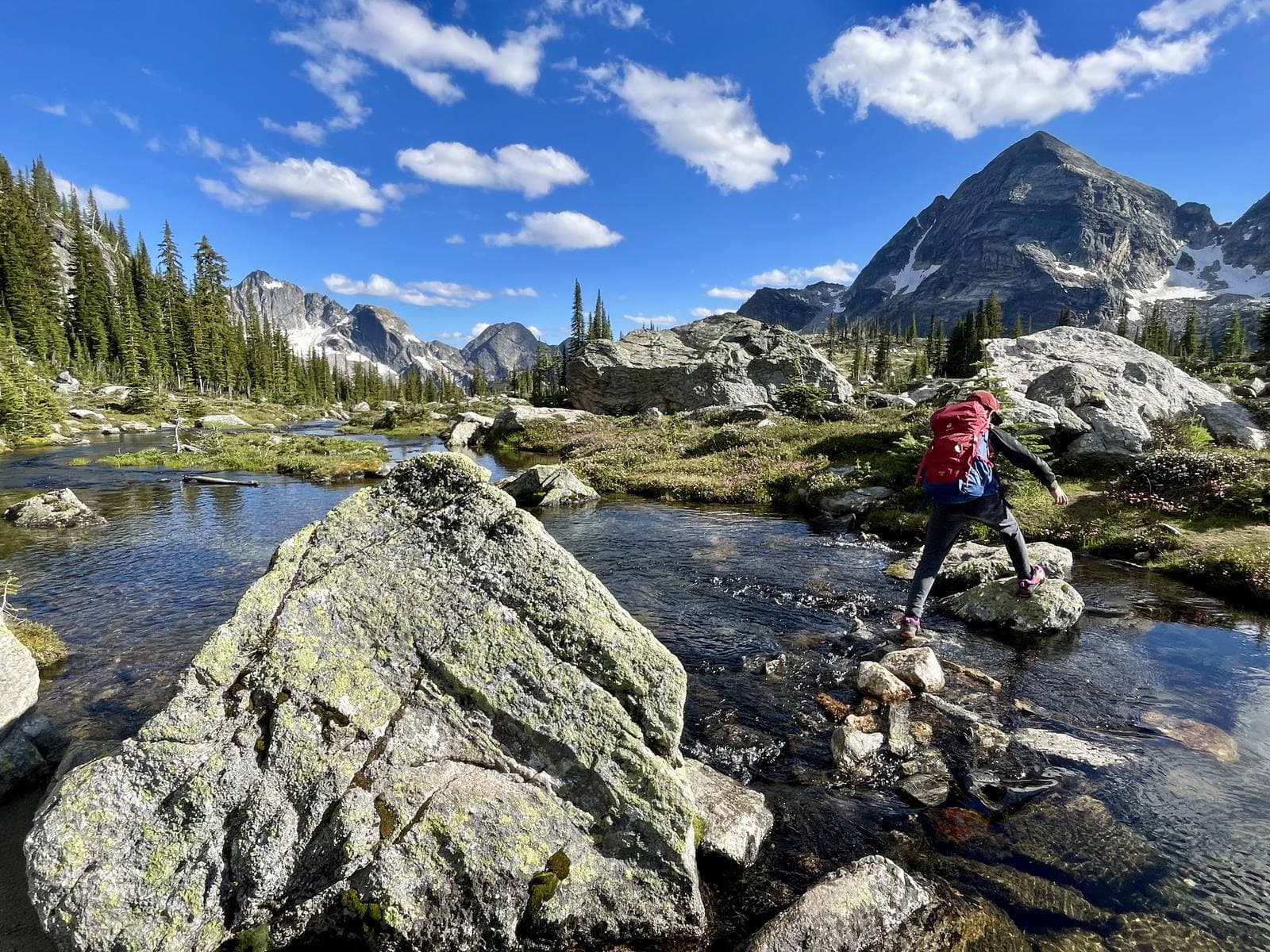 A kid with a red backpack rock-hopping across a clear mountain creek, with alpine meadows, boulders and a rocky peak behind