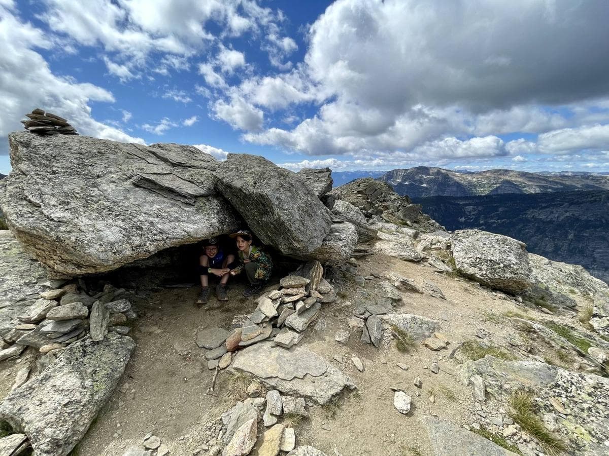 Two kids crouched in a natural shelter under a big rock overhang on a mountain ridge, with a stone cairn on top and mountains behind