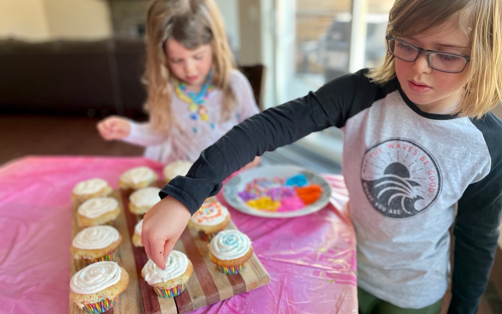 Two kids decorating a tray of homemade cupcakes with white icing at a table covered in a pink party tablecloth, a plate of coloured sprinkles between them