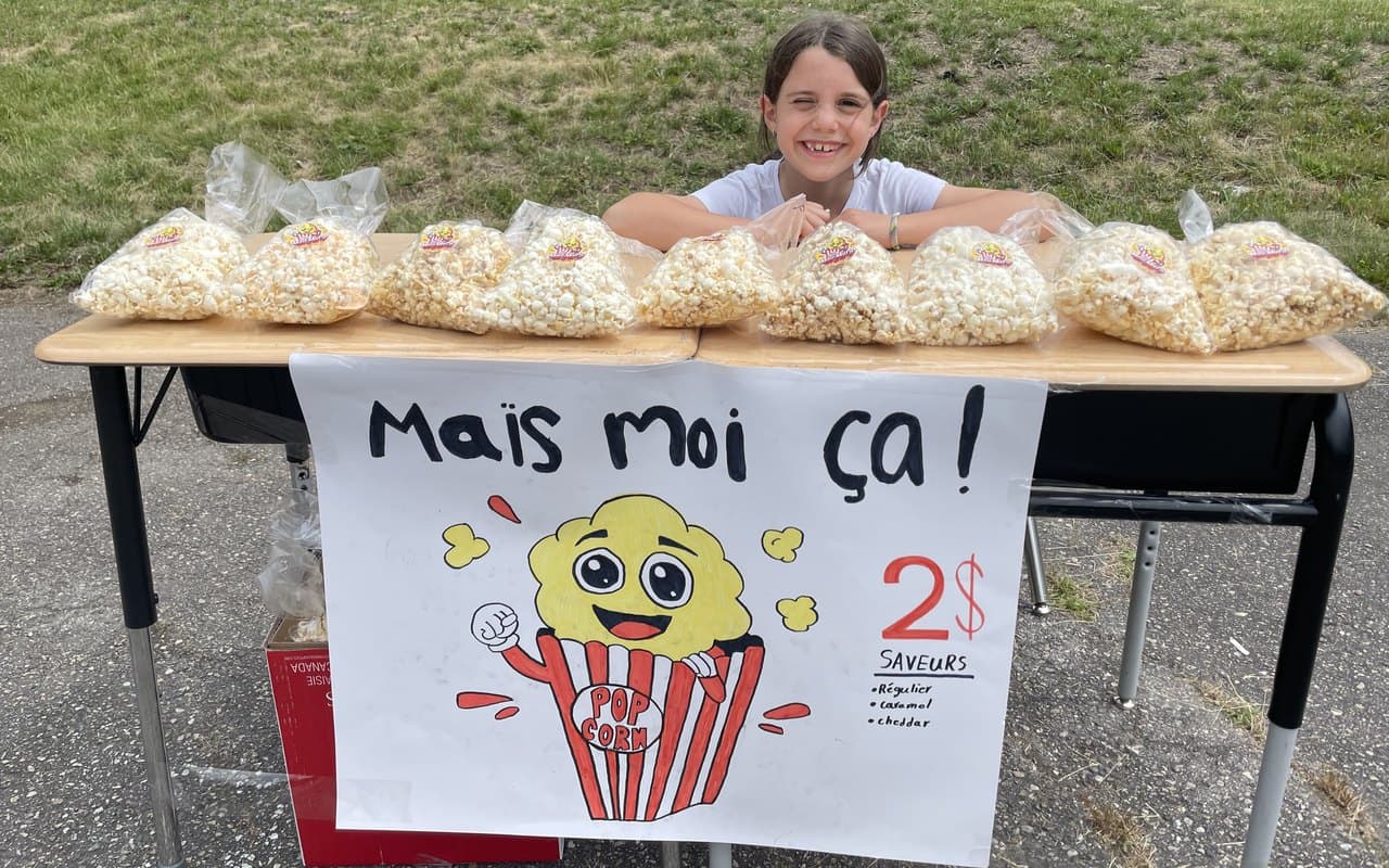 A smiling girl running her own popcorn stand outdoors, with rows of brand-labeled bags she made and a hand-painted sign listing the price and flavors