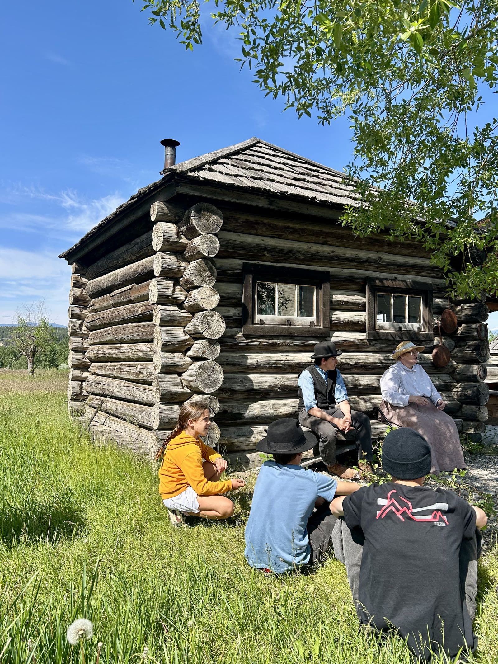 Kids sitting in the grass outside a pioneer-era log cabin while people in period costume sit on the porch, a living-history scene in action
