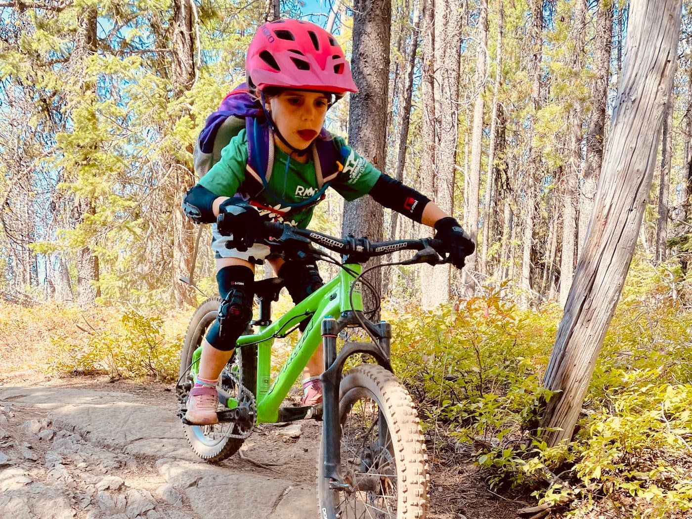 Kid in a pink helmet and knee pads riding a mountain bike over a rocky technical section on a forest trail