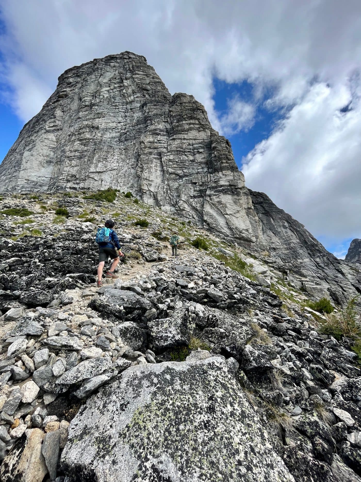 A kid with a backpack scrambling up a scree slope toward a massive cliff, another hiker visible on the trail ahead