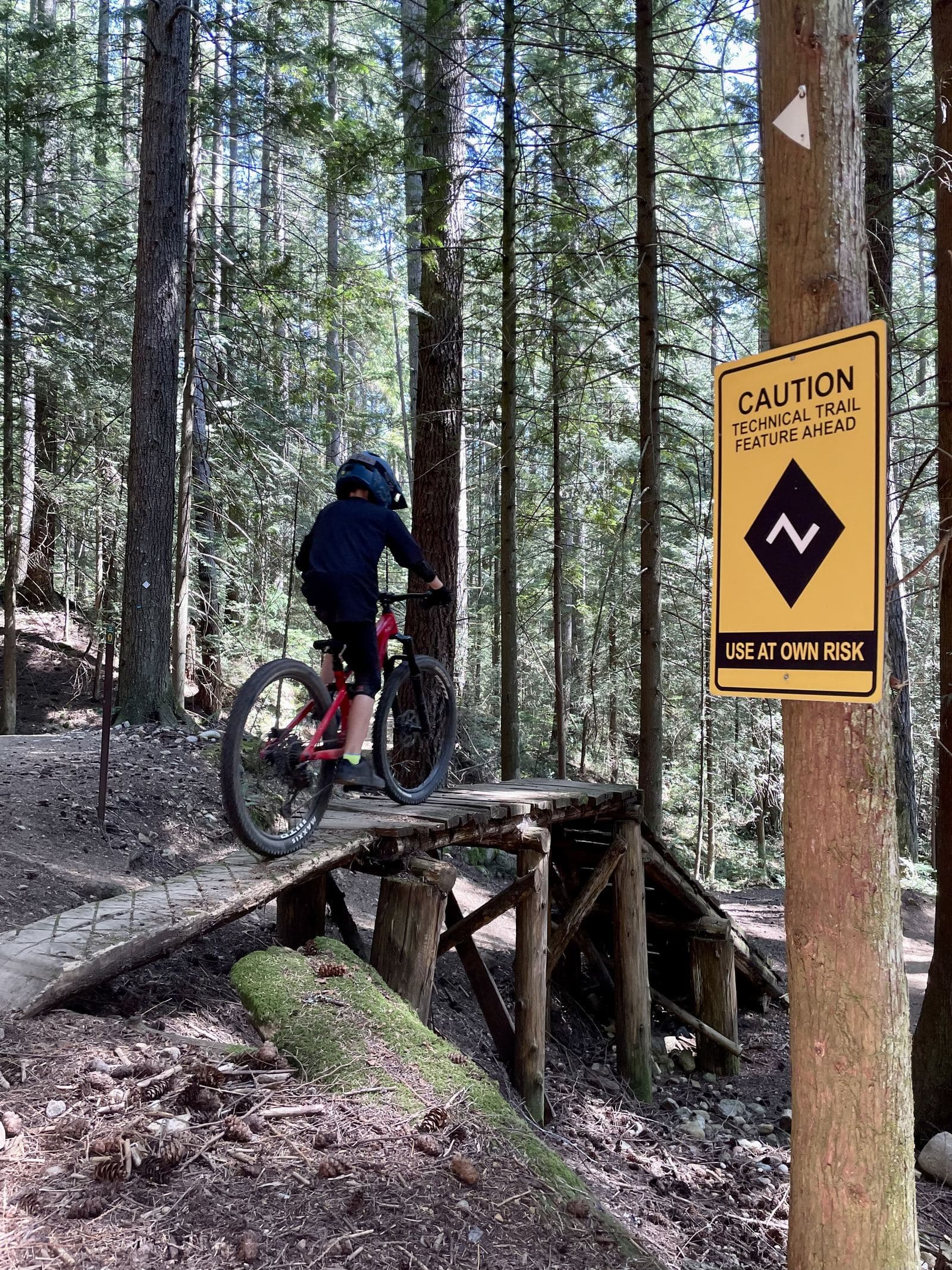 Kid on a mountain bike crossing a wooden trail feature next to a yellow caution sign reading USE AT OWN RISK