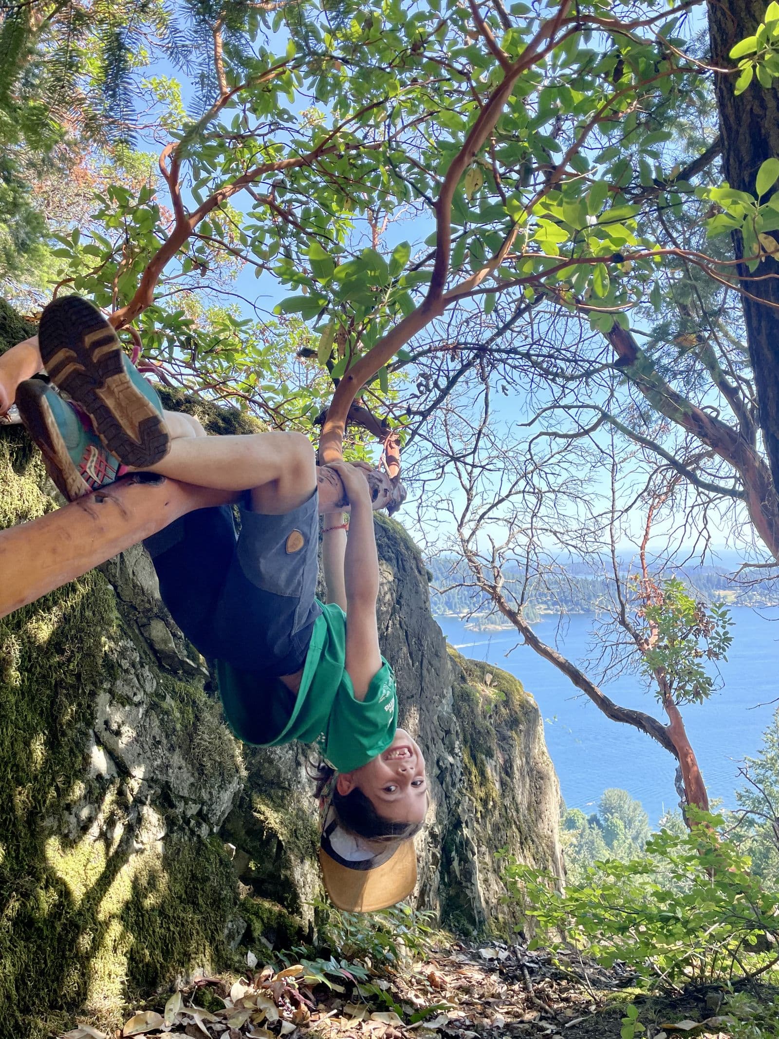 Kid hanging upside down from a tree branch on a cliff edge above the ocean, grinning, the joyful side of risky play