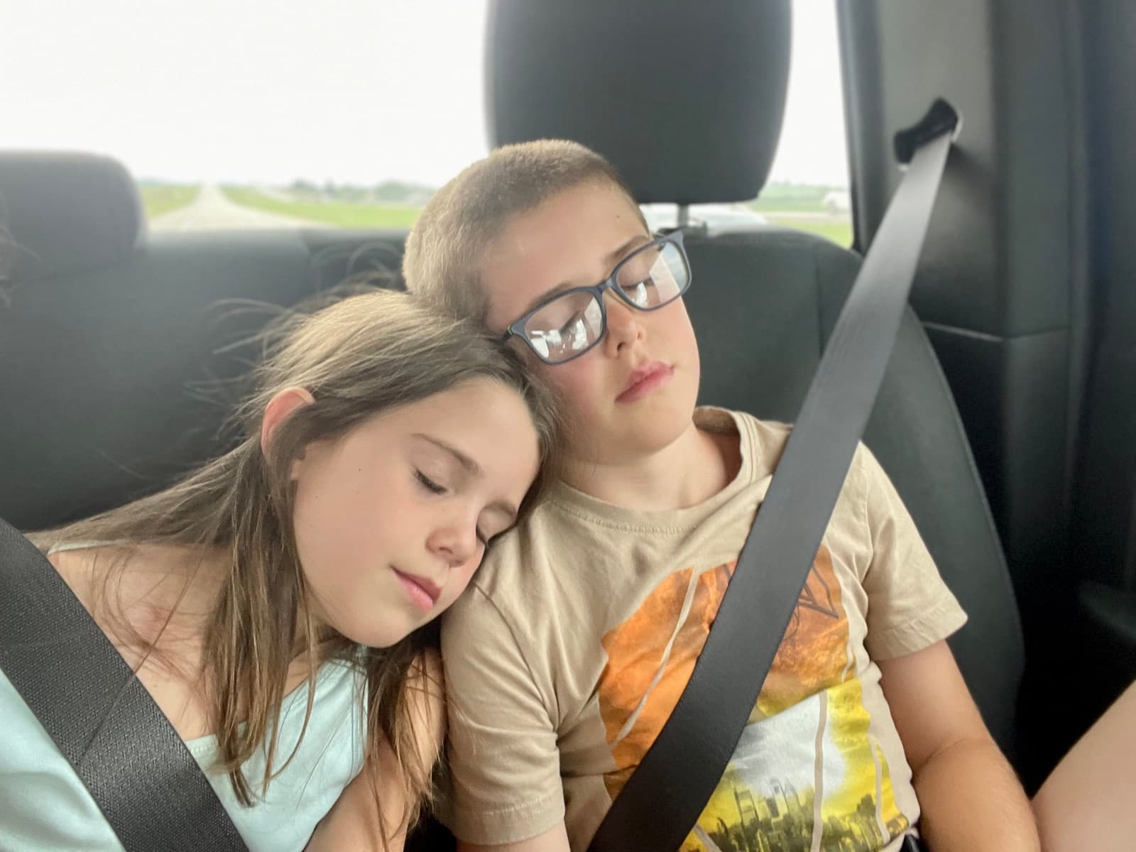Two kids asleep against each other in the back seat on a long drive, seatbelts on, the highway stretching out through the windshield
