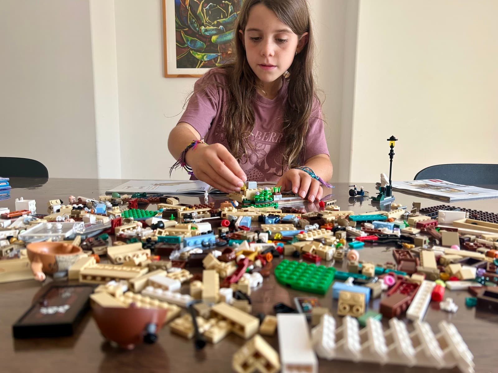 Julia at a dining table deep in a lego build, surrounded by piles of pieces, completely absorbed in the project — no screen in sight