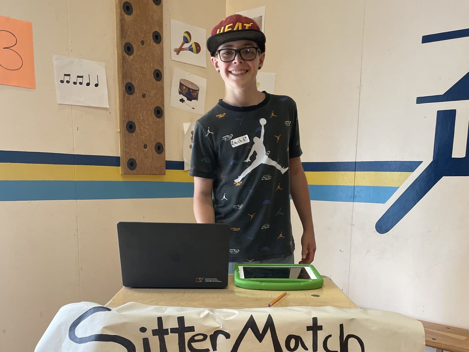 A tween standing behind his pitch table presenting an invented app called SitterMatch, with a hand-drawn banner, a real business pitch