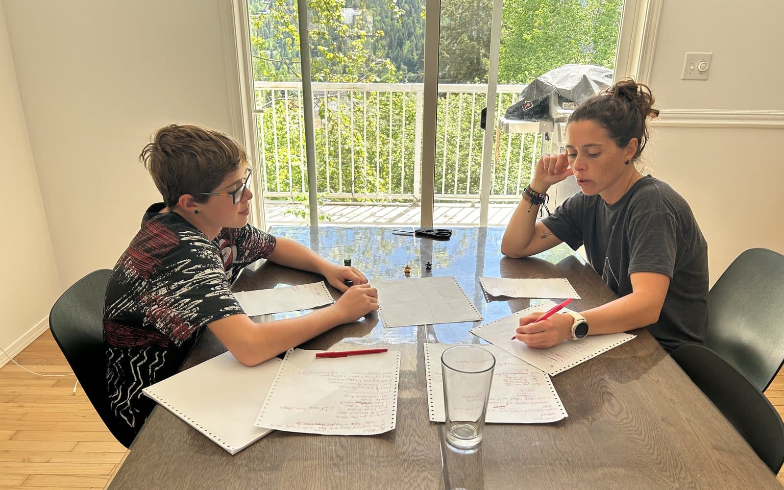 A mother and her son sitting across a dining table at home, both working on paper together in the morning light