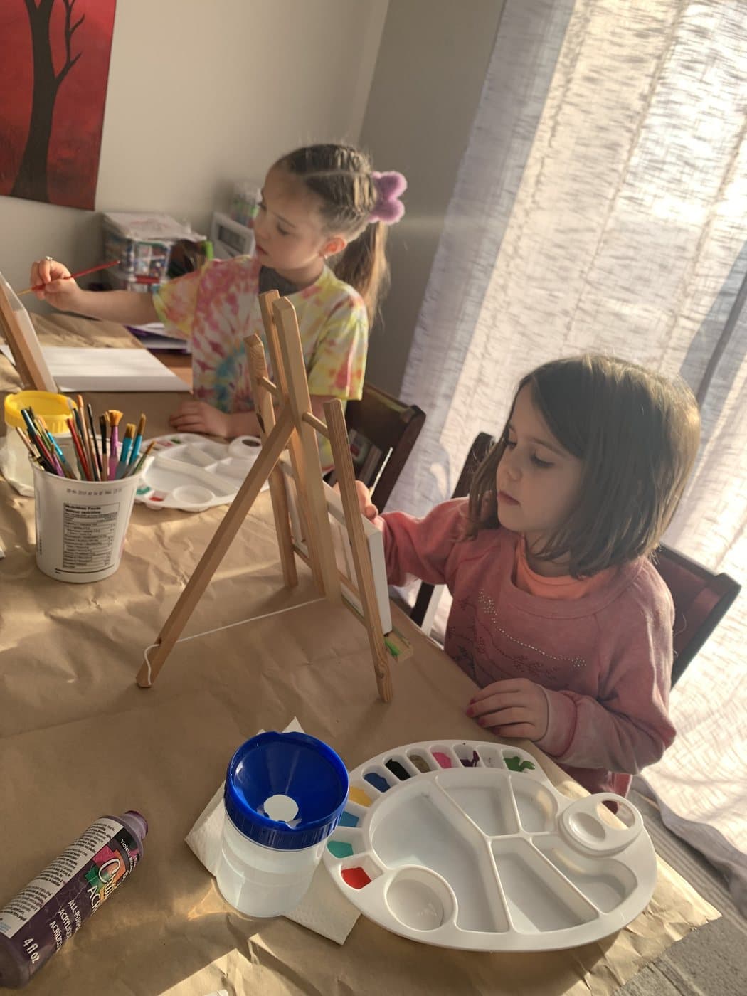 Two children painting at an easel and table at home, brushes and a paint palette in front of them