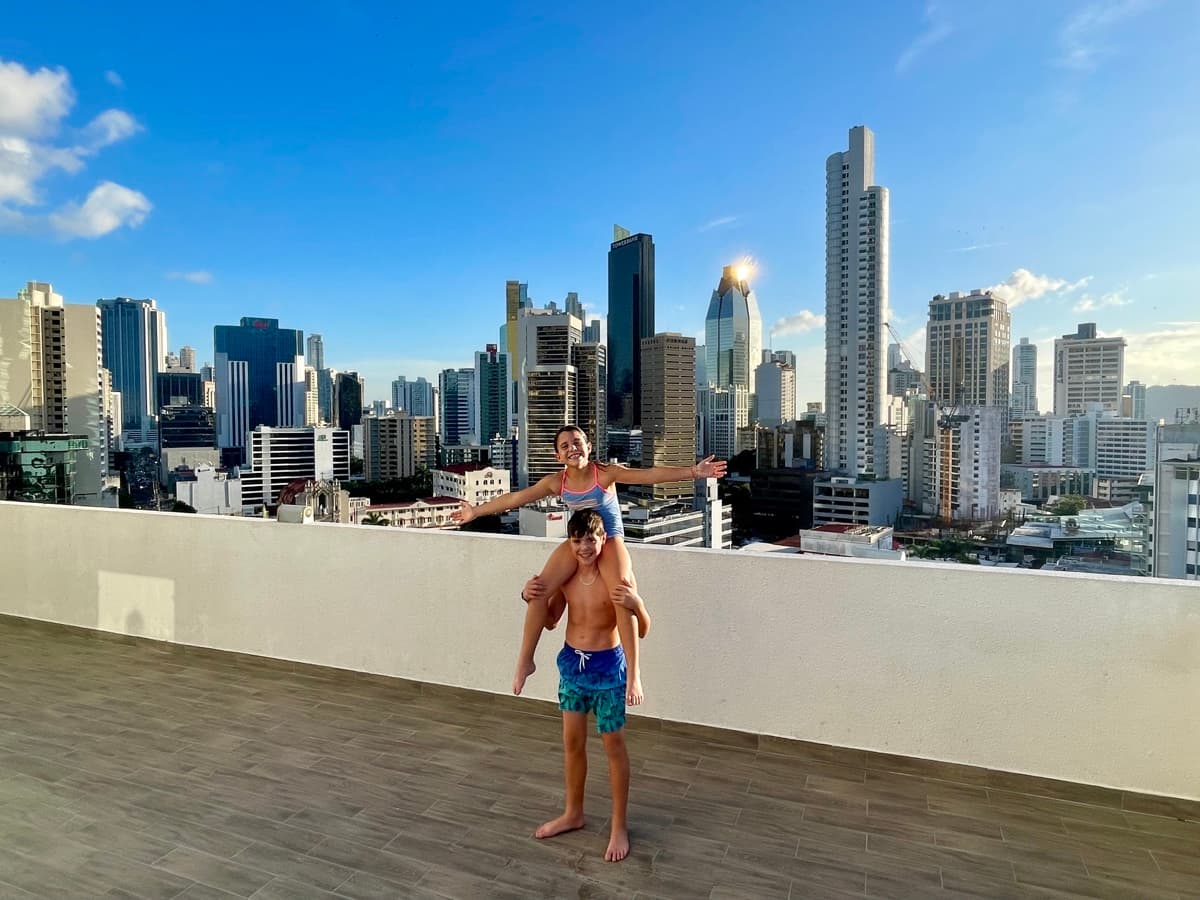 Julia on Zach’s shoulders with arms wide open on a rooftop with the Panama City skyline behind them