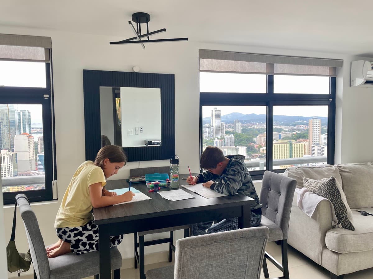 Two kids working at a table in a bright apartment with a city skyline and mountains through the windows