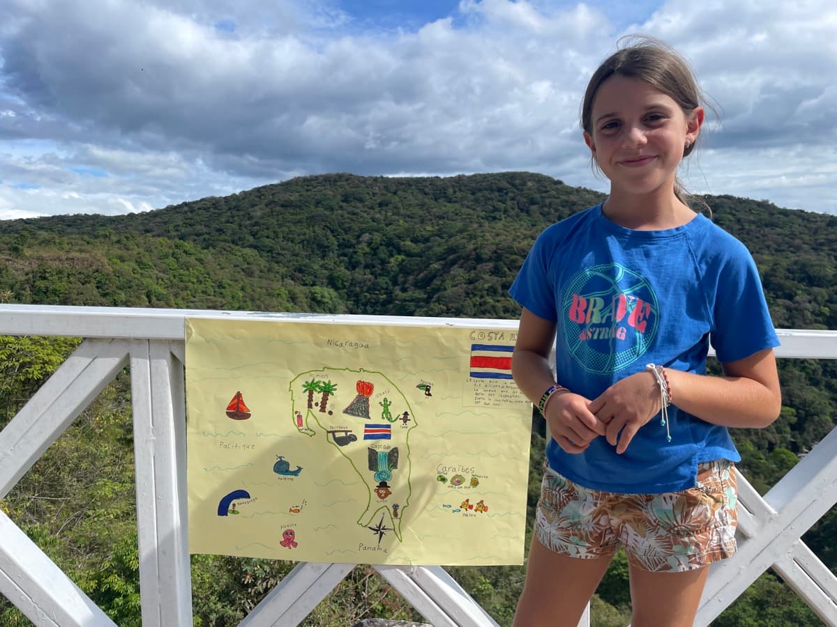 My daughter proudly holding up her hand-drawn Costa Rica poster with lush green hills behind her