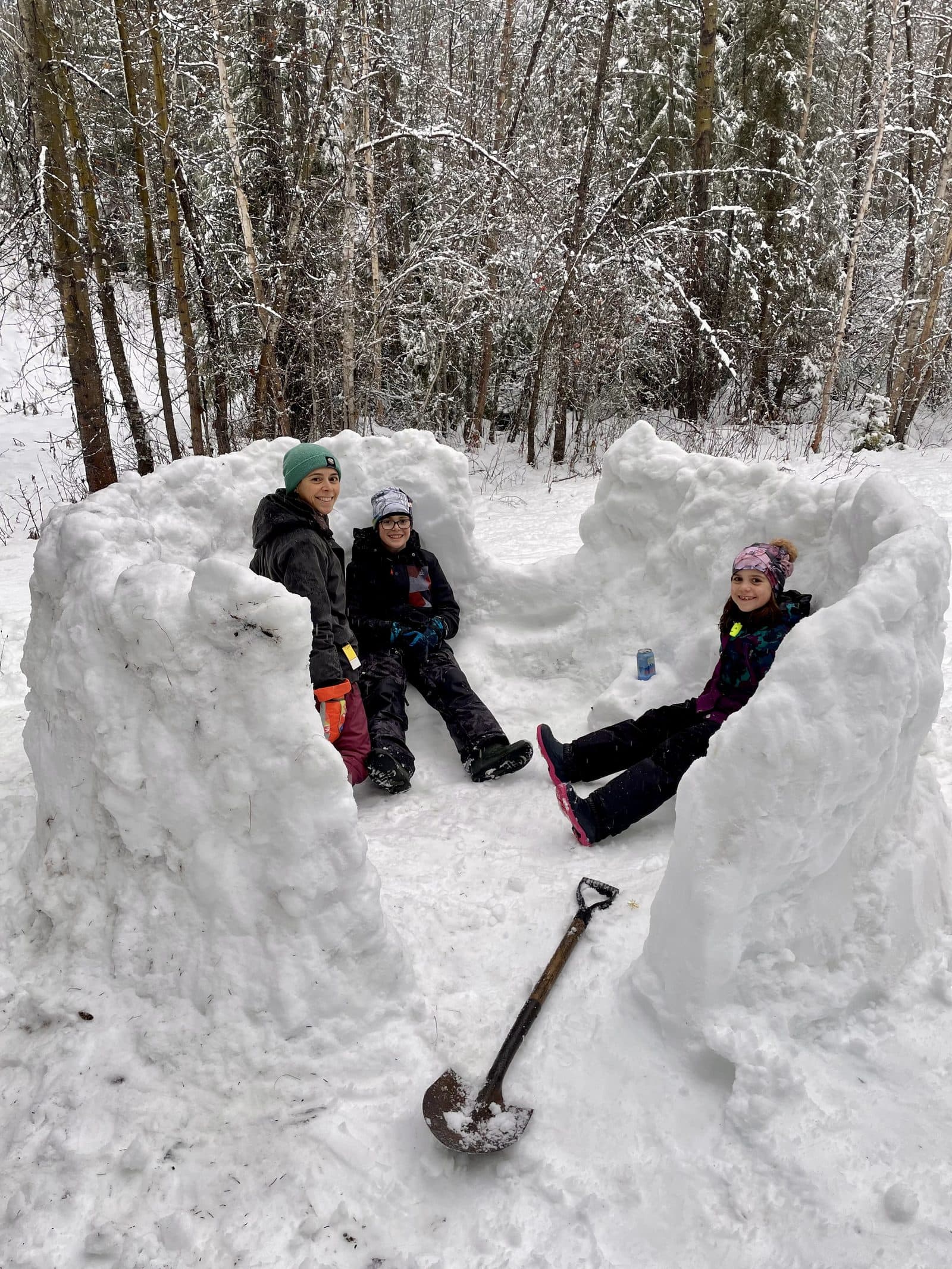 Mom and two kids of different ages sitting inside the snow fort they built together in the woods, shovel in the foreground, real winter engineering at family scale